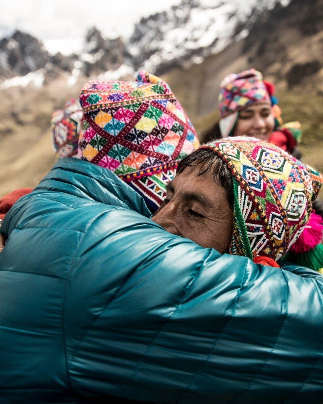 Some connections happen fast, especially at altitude. Two people wearing colorful hand-embroidered Andean chullo hats share a warm embrace on the salkantay hike, with snow-dusted mountain peaks rising in the background and a smiling woman looking on