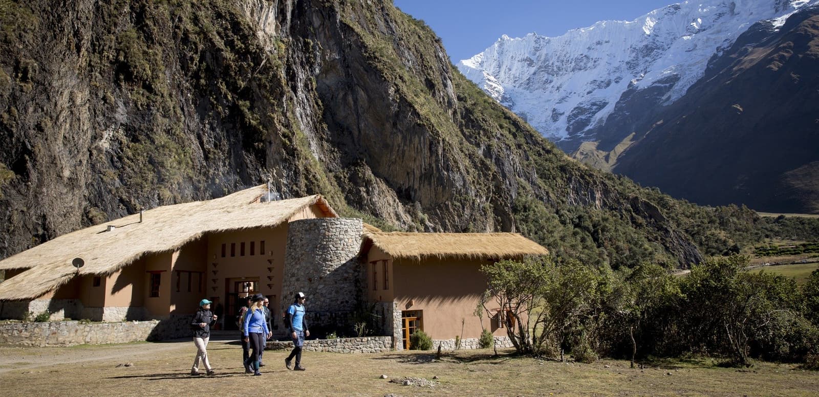 Due notti qui, un ghiacciaio in vista, e la fatica che diventa ricordo. Tre escursionisti si allontanano da un lodge in pietra e adobe con tetto di paglia durante l'escursione al Salkantay, con ripide pareti rocciose e una cima innevata sullo sfondo