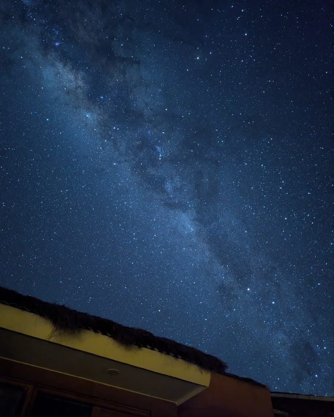 No light pollution, no problem. The galaxy shows up every night. The Milky Way arching across a deep blue starry sky above the dark silhouette of a thatched-roof lodge on the Salkantay hike route in the Peruvian Andes