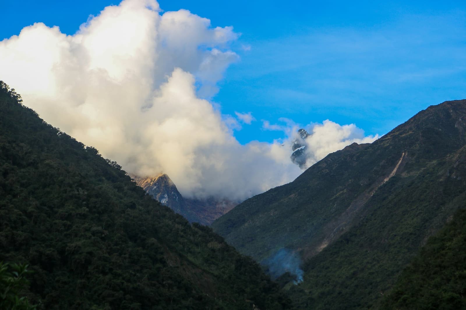 As nuvens não passam por aqui. Elas ficam. Vale profundo e coberto de floresta no trek Salkantay de 5 dias, com picos rochosos parcialmente encobertos por nuvens volumosas e um fio de fumaça subindo do fundo do vale
