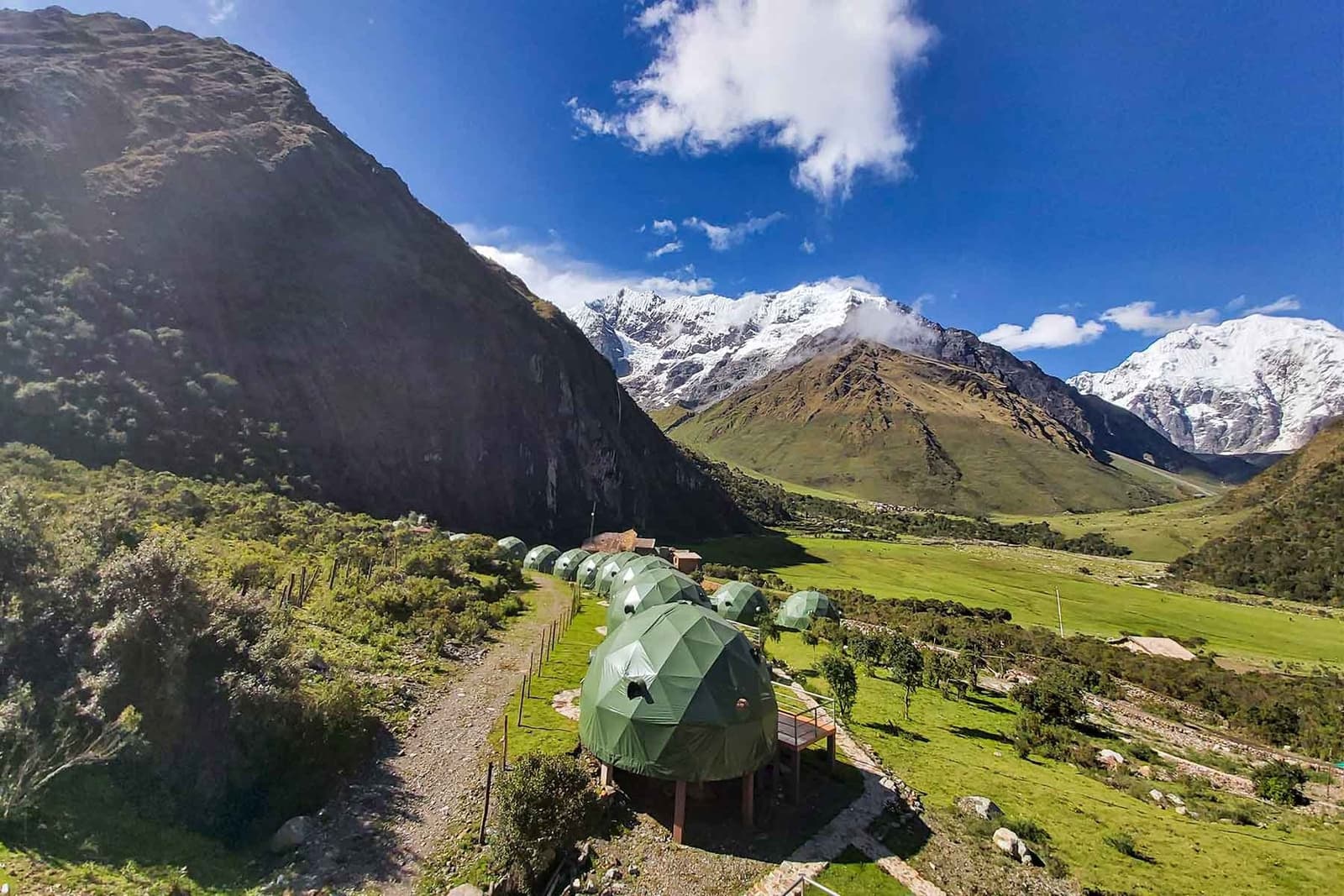 Dormir de frente para os nevados, sem abrir mão de uma cama de verdade. Uma fileira de domos geodésicos verdes em plataformas de madeira elevadas ao longo de um caminho na encosta, com um amplo vale verde e picos andinos cobertos de neve ao fundo na trilha Salkantay de 5 dias