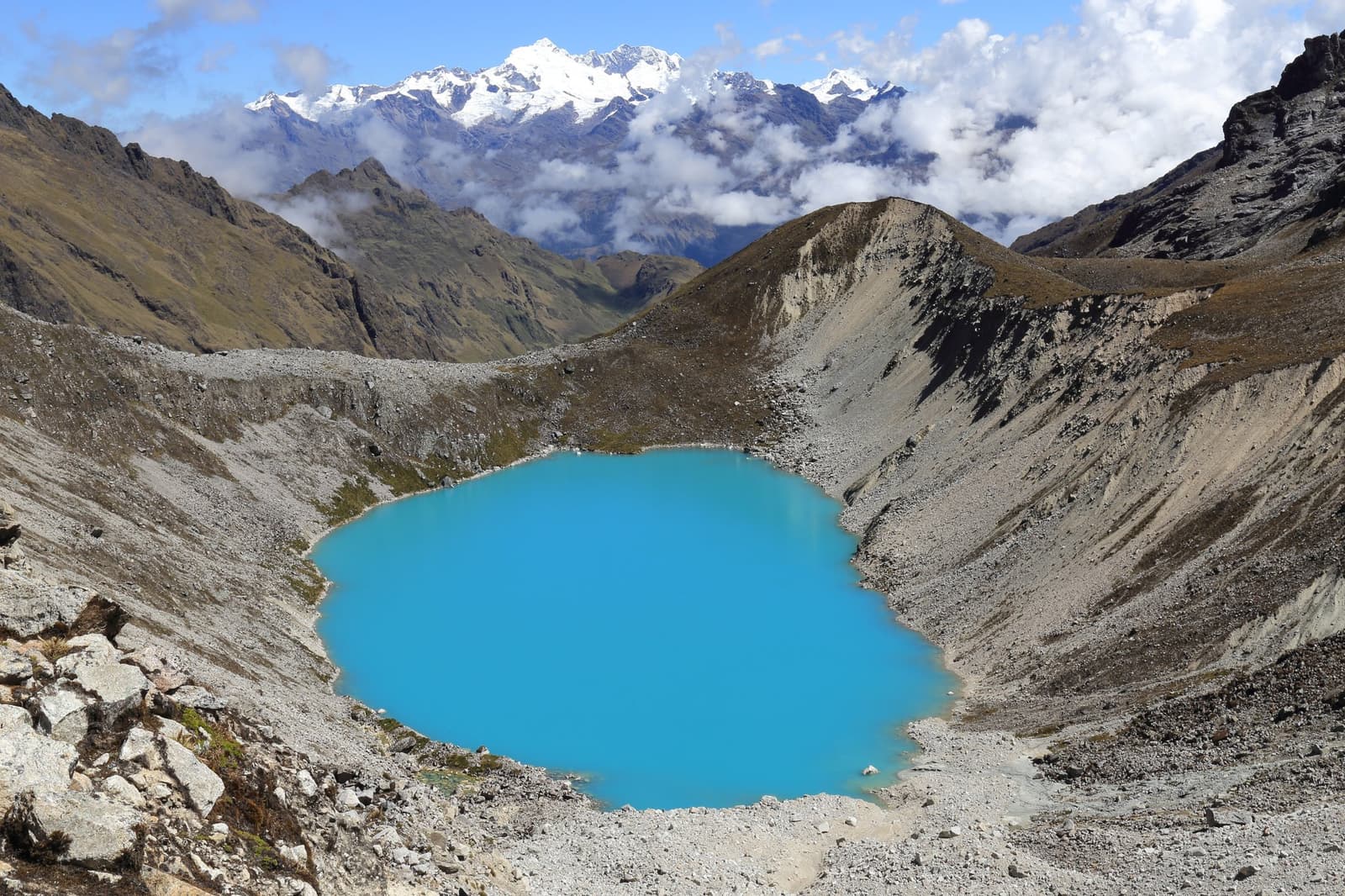 Esse azul é exatamente assim. Sem filtro, sem exagero. Um lago glacial de turquesa intenso encaixado em um circo rochoso de alta altitude no trek Salkantay de 5 dias, com picos andinos cobertos de neve atrás de nuvens baixas ao fundo