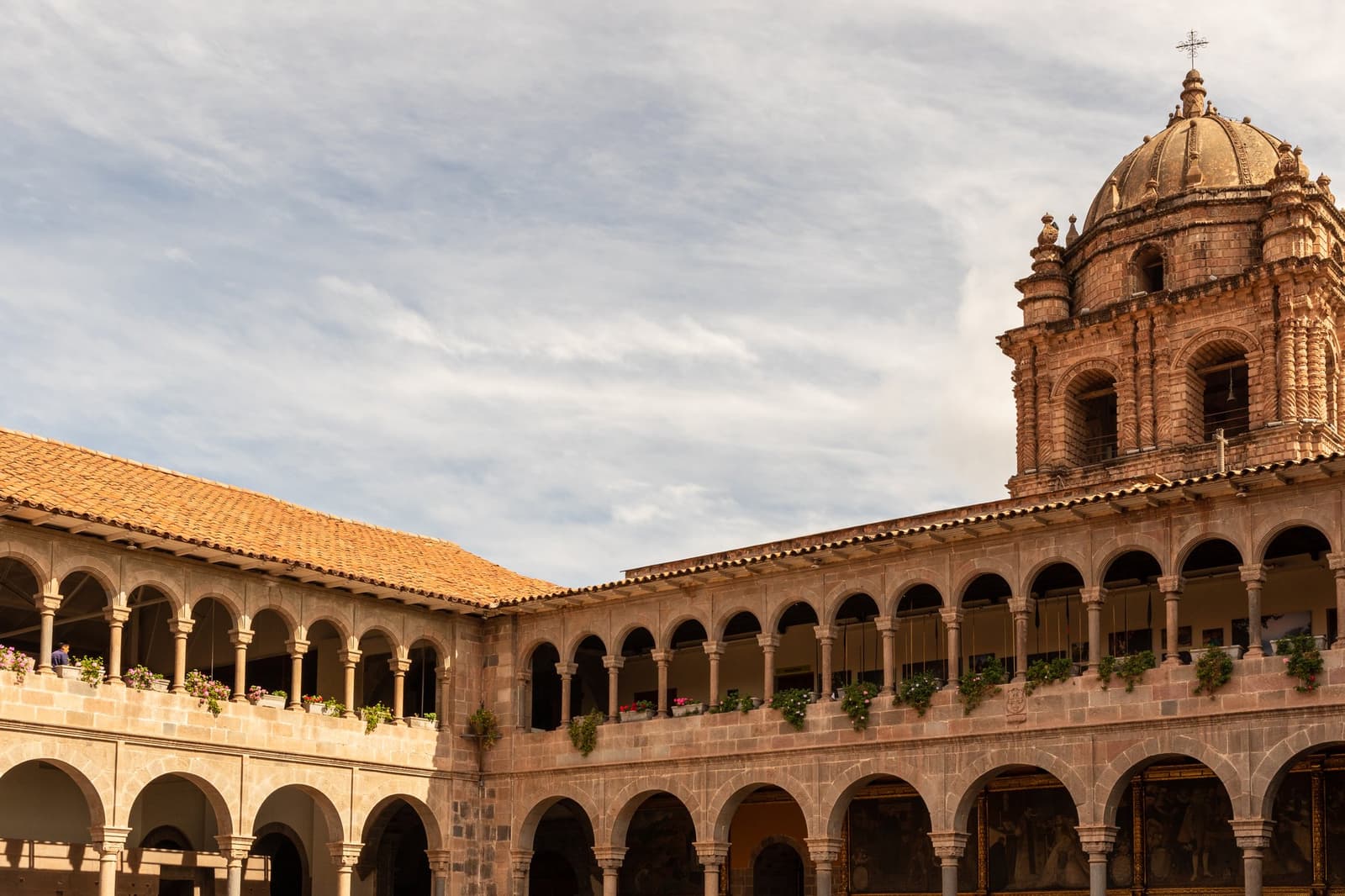 Cusco define o ritmo: caminhe devagar, olhe para cima e deixe a altitude fazer seu papel. Claustro de pedra com dois níveis de colunatas em arco, vasos de flores e uma cúpula barroca de igreja se erguendo acima dos telhados de terracota em Cusco, Peru