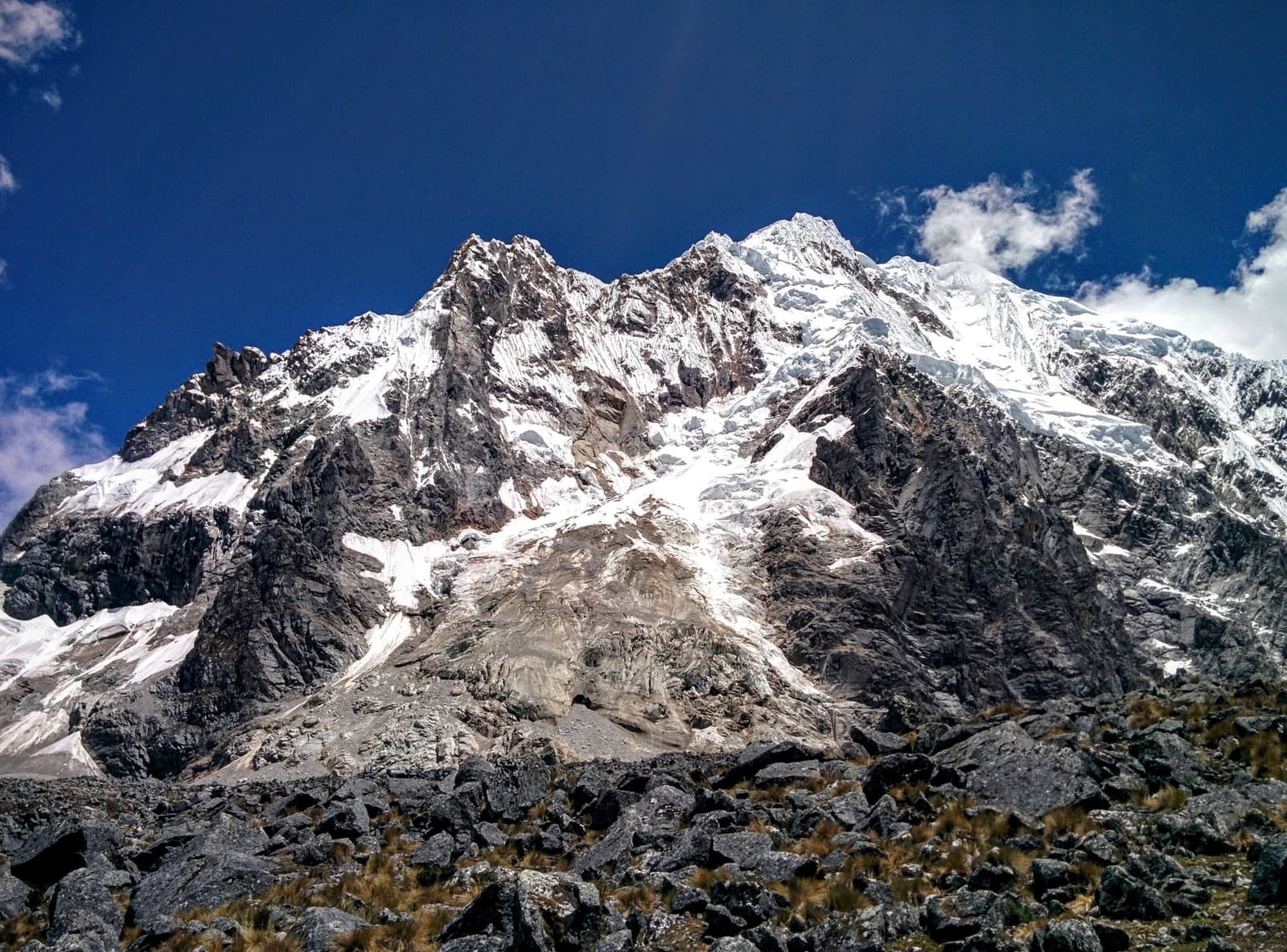 De perto, a geleira deixa bem claro quem manda aqui. Pico dentado coberto de neve com faces rochosas expostas e um campo de morena glacial em primeiro plano, visto na rota do trek Salkantay ao Machu Picchu sob um céu azul profundo