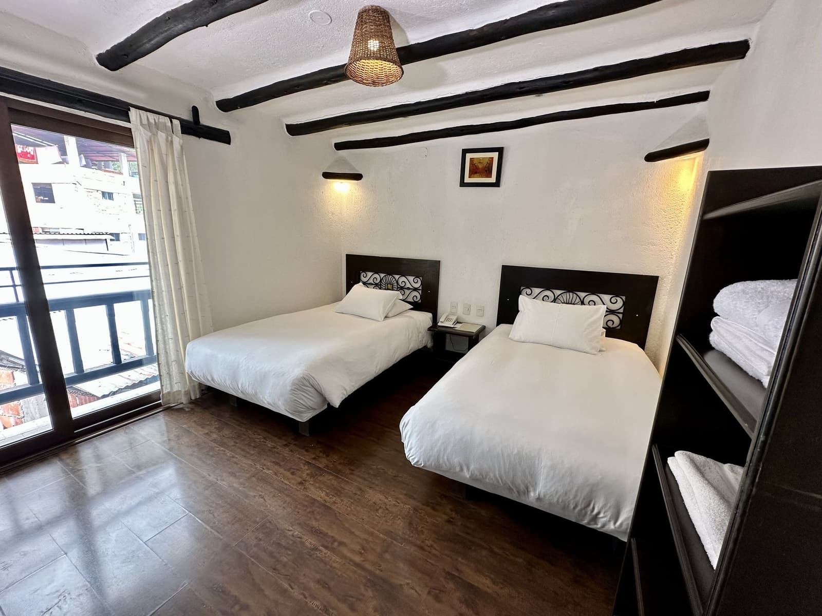 Schone lakens en een balkon, precies genoeg comfort voor de volgende ochtend. Hotelkamer met twee eenpersoonsbedden, witte lakens, donkere houten dakbalken, een rieten hanglamp en een balkon met uitzicht op een stadsstraat