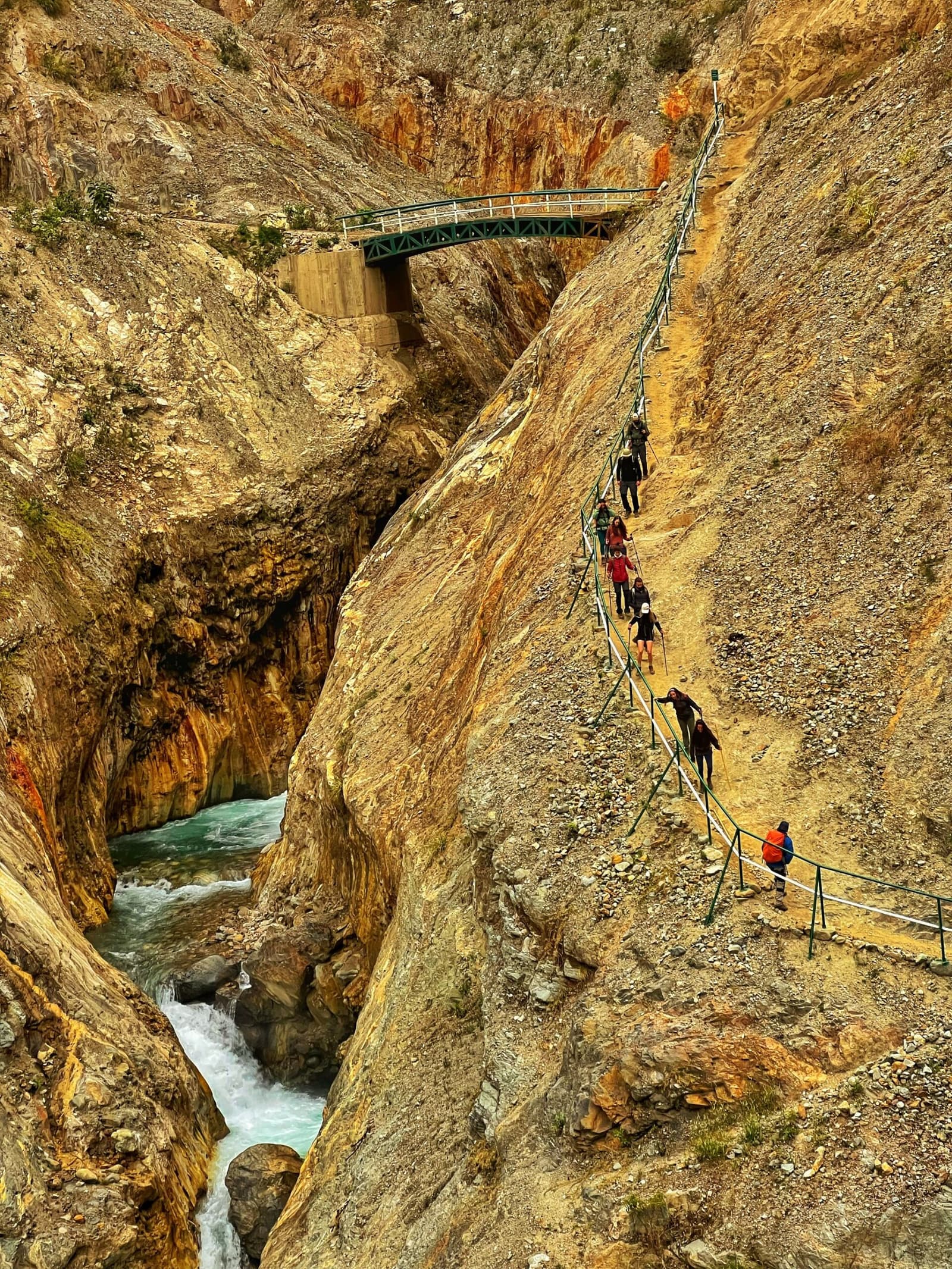 Passo a passo, o cânion aperta e o rio vai ficando mais alto. Um grupo de caminhantes desce uma trilha íngreme com corrimões metálicos esculpida em paredes de cânion cor de ocre durante o trek Salkantay a Machu Picchu, com um rio turquesa correndo pela garganta lá embaixo e uma ponte metálica visível ao fundo