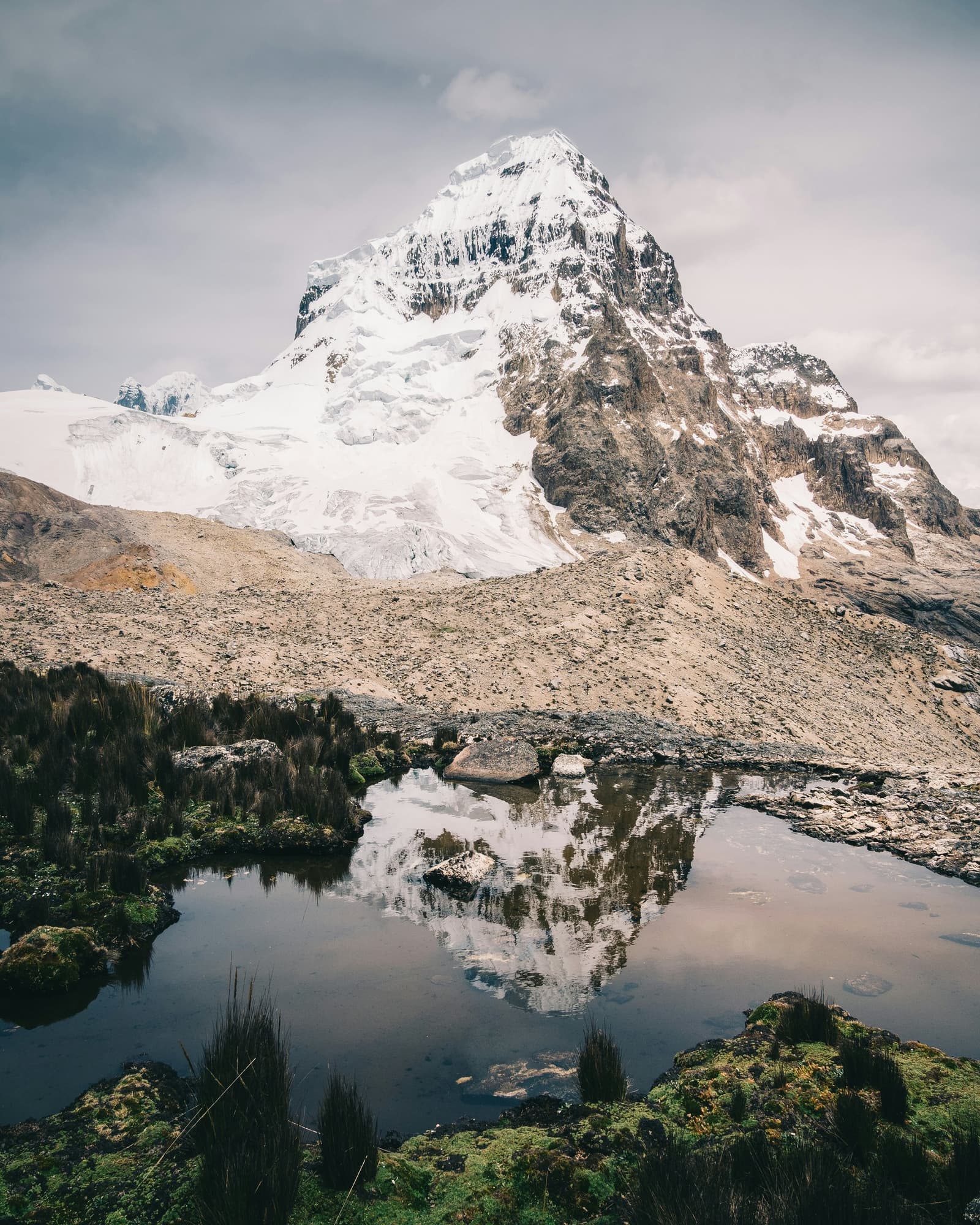 Água parada a quase 4.000 metros tem uma honestidade que a montanha aprecia. Um pico rochoso coberto de neve se ergue acima de um lago glacial de altitude na trilha Santa Cruz, com seu reflexo perfeitamente capturado na água escura e calma, e pedras cobertas de musgo e gramíneas em tufos no primeiro plano