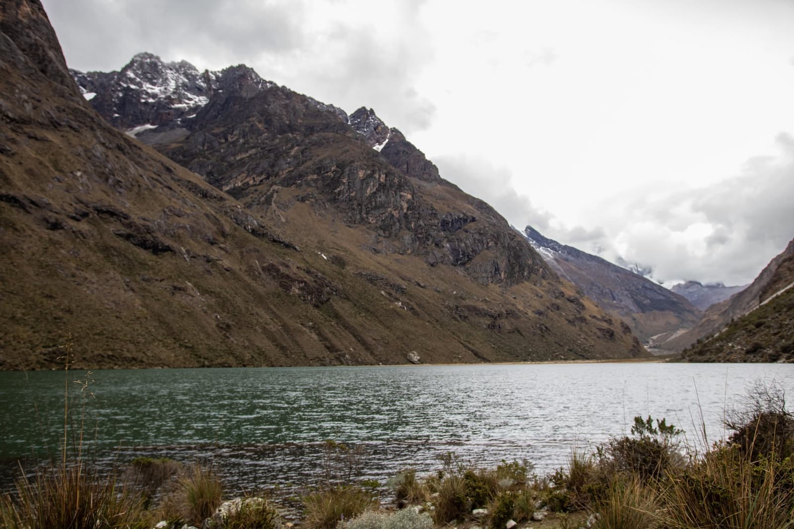 Cold water, big walls, and clouds that can't quite make up their minds. A green glacial lake on the santa cruz trek reflecting overcast skies, surrounded by steep rocky Andean slopes with snow-dusted peaks disappearing into low cloud