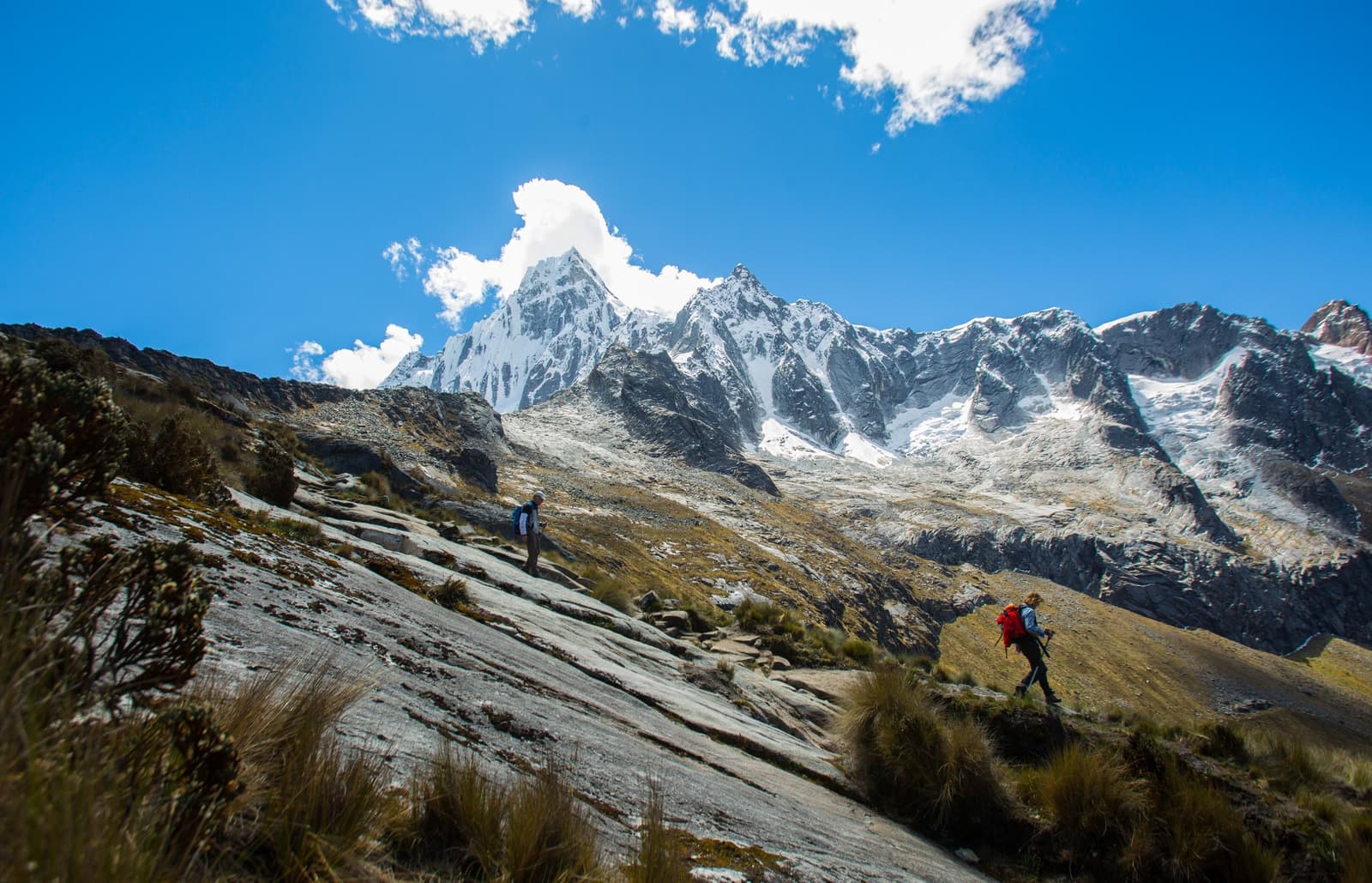 The mountains here don't ease you in, they show up all at once. Two trekkers with backpacks climbing a rocky granite slope on the santa cruz trek, with heavily glaciated peaks of the Cordillera Blanca rising sharply under a deep blue sky behind them