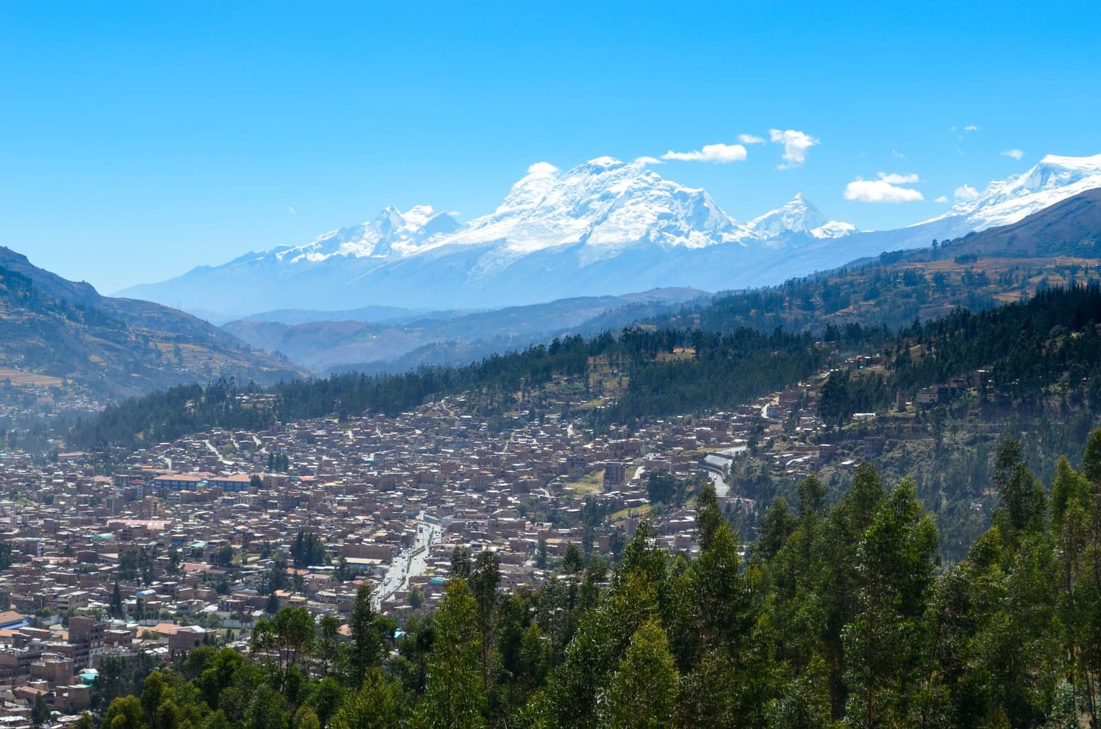 Huaraz ocupa seu vale com calma, e as montanhas nevadas cuidam do horizonte. Vista panorâmica aérea sobre os telhados densos da cidade de Huaraz, encaixada em um vale andino, com os picos nevados da Cordilheira Branca se erguendo por trás de encostas florestadas sob um céu azul límpido.