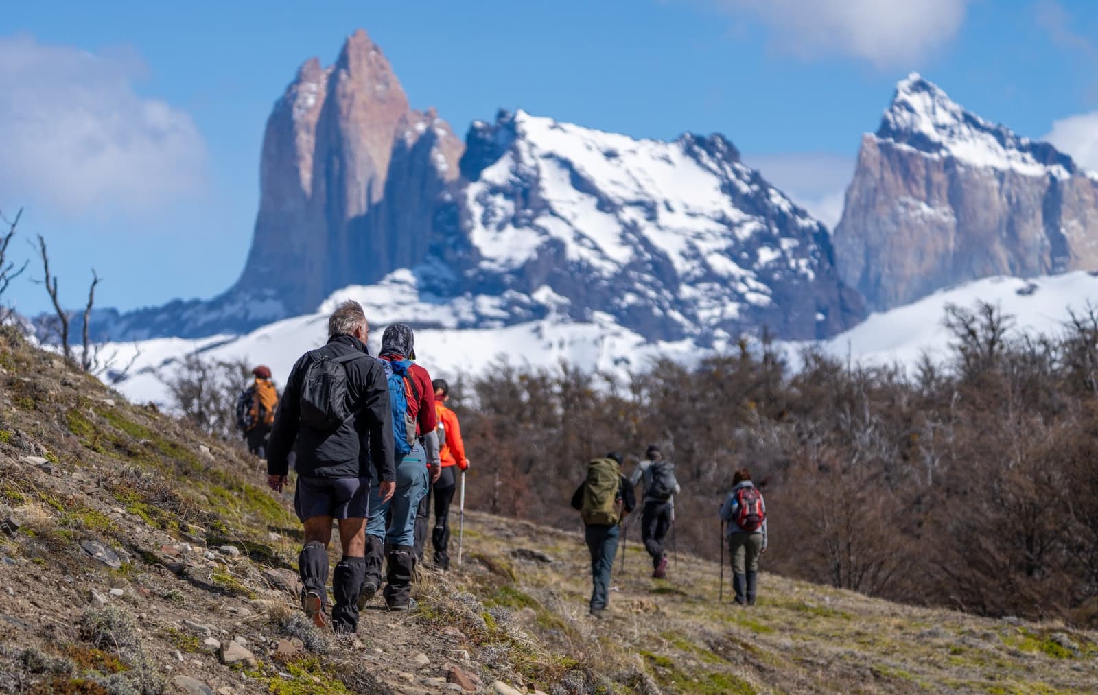 Six people, one trail, zero guide, plenty of granite. A group of hikers with backpacks and trekking poles climbs a rocky hillside trail on a self-guided W Trek, with jagged snow-capped granite peaks rising sharply against a blue sky in the background.