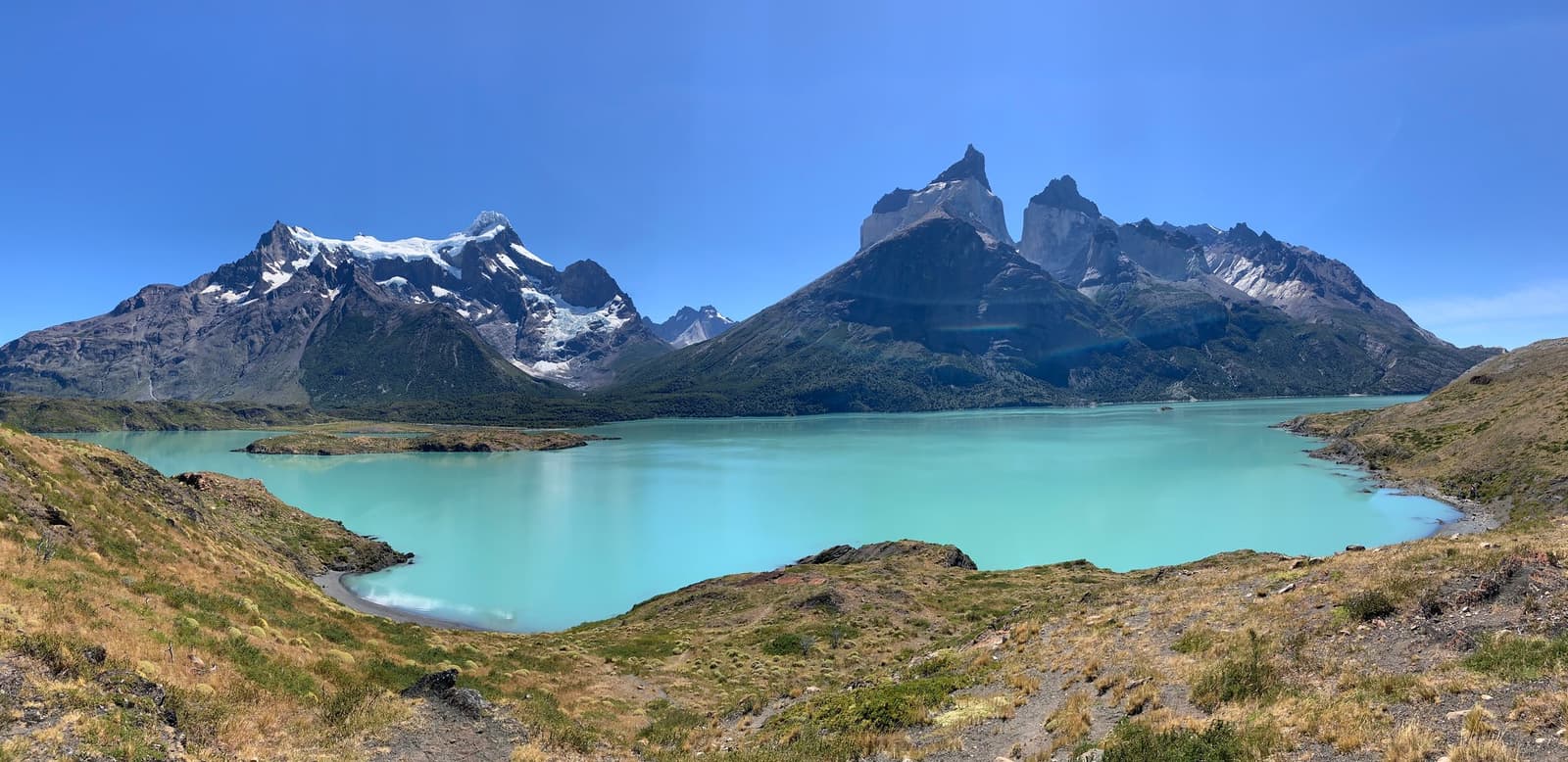 That milky turquoise colour comes from glacial flour. The lake earns it. Panoramic view of a glacial turquoise lake reflecting the Cuernos del Paine granite peaks under a clear blue sky, seen from a grassy hillside on the self-guided W Trek in Torres del Paine National Park