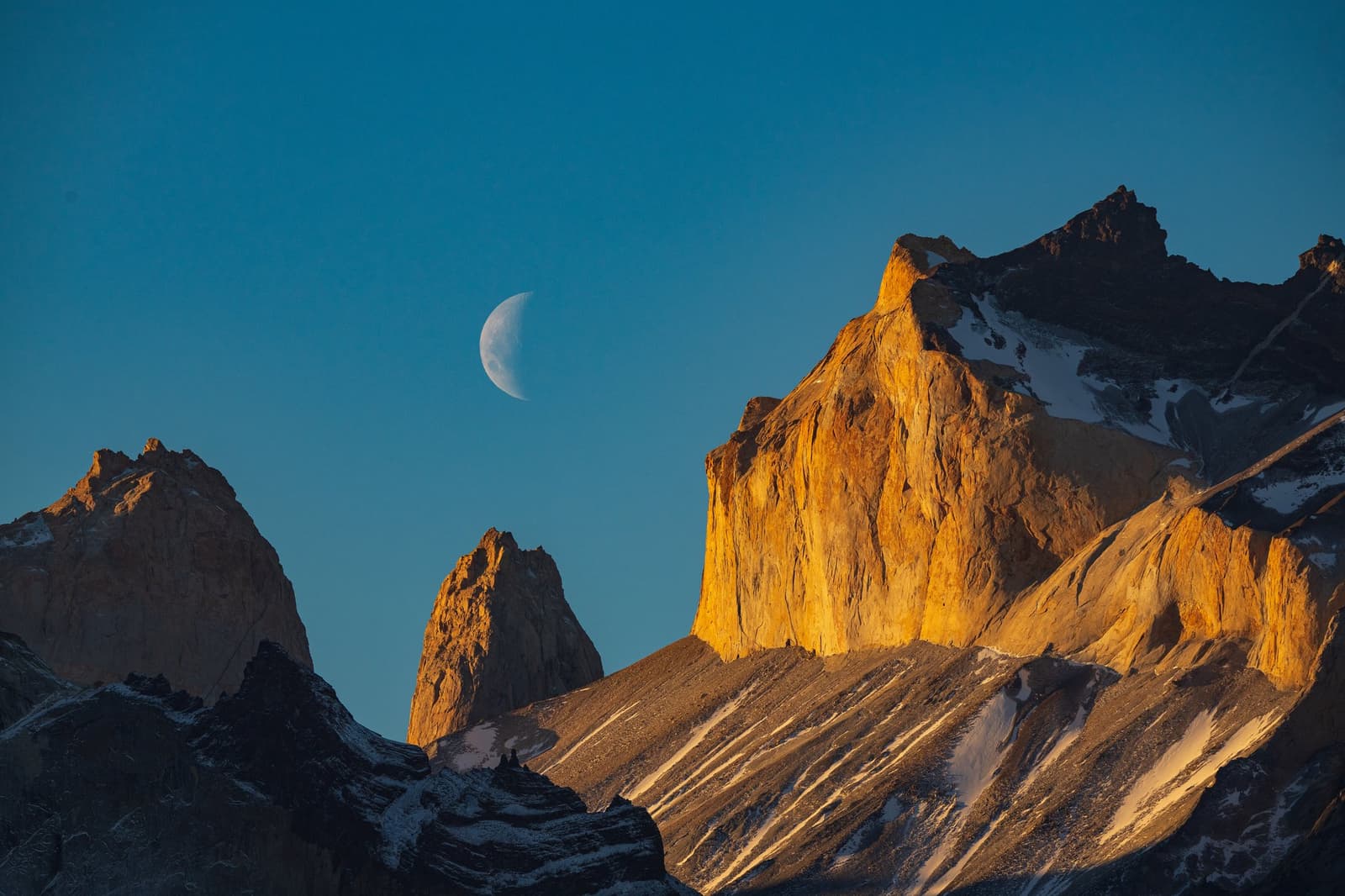 Moon still up, rock already on fire. Patagonia keeps its own schedule. Golden alpenglow lights the jagged granite peaks of the Cuernos del Paine on the self-guided W Trek, with a crescent moon hanging in the deep blue sky above the snow-dusted ridgeline.
