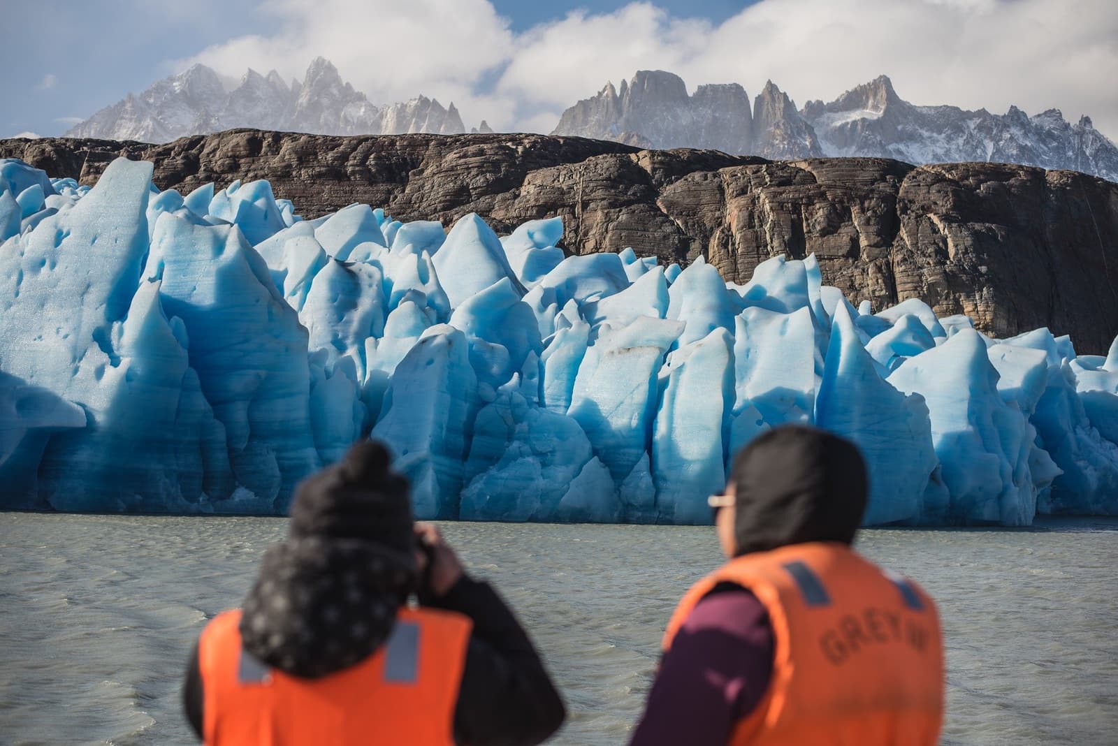 That blue is ancient compressed ice, and yes, your drink was chilled with a piece of it. Two travelers in orange life vests photographing the jagged blue ice wall of Grey Glacier from a boat on the short W trek, with snow-dusted granite peaks rising behind
