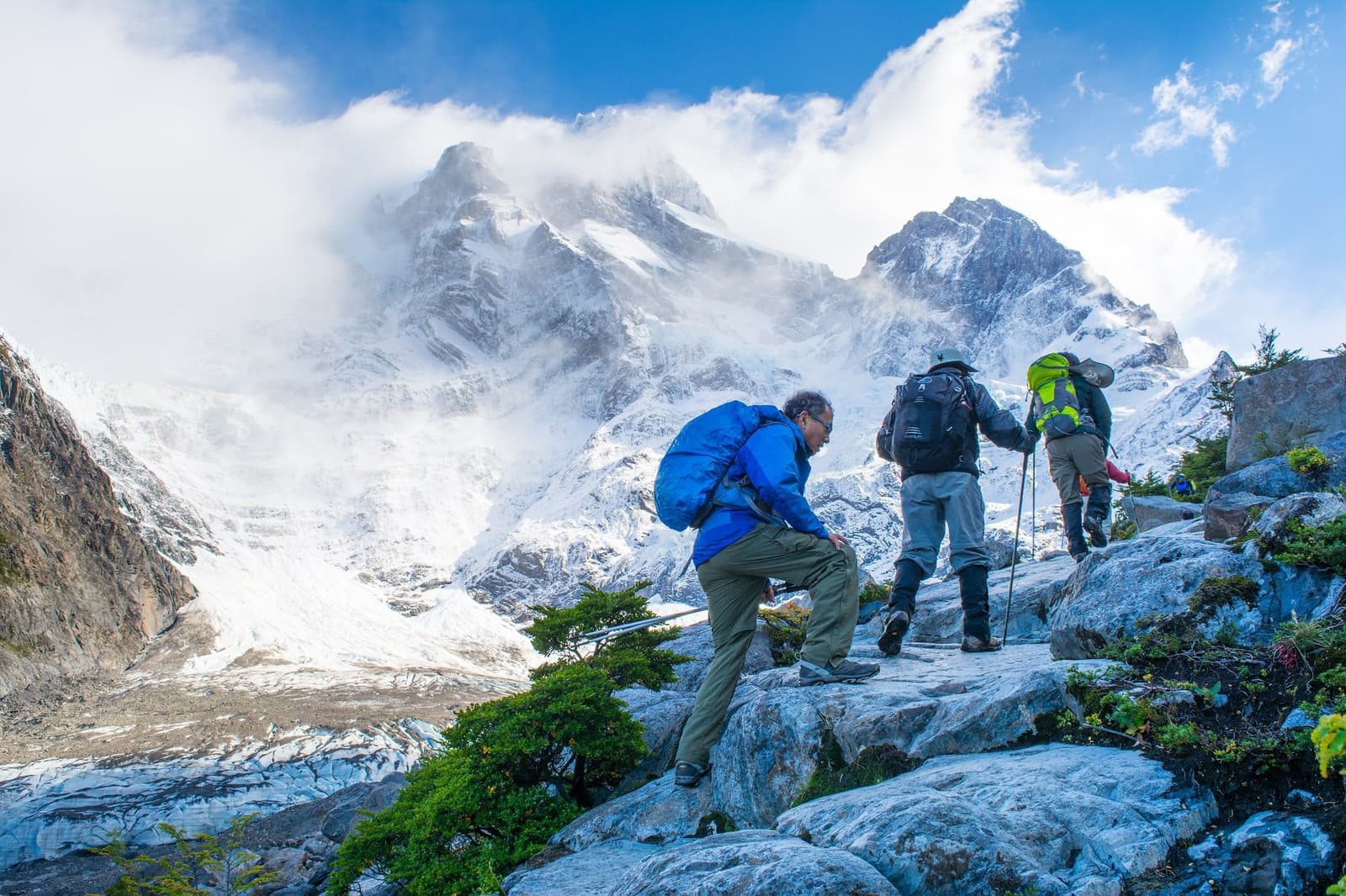 Legs burning, scenery not helping you slow down. Three hikers with backpacks scrambling up a rocky ridge on the short w trek, with a snow-covered mountain peak wrapped in clouds and a glacier visible below in the valley