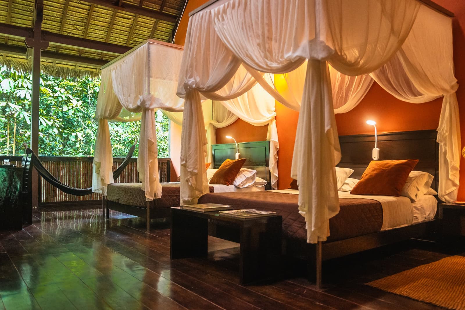 Jungle sounds come free with every room. The hammock is just a bonus. Twin four-poster beds with white draped canopies and terracotta walls in a Tambopata Research Center Peru lodge room, with dark hardwood floors, warm reading lights, and an open veranda overlooking dense rainforest with a hammock visible outside