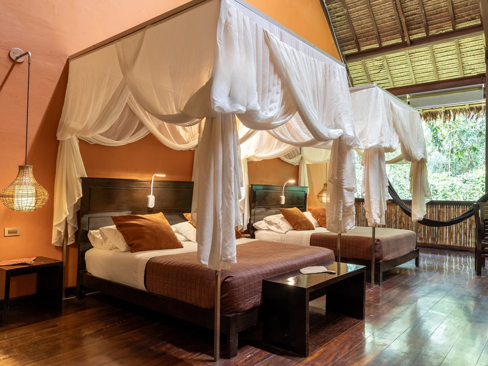 Fall asleep to the forest, wake up to it too. Twin four-poster beds with white mosquito net canopies in a Tambopata Research Center Peru lodge room, with warm terracotta walls, dark hardwood floors, rattan pendant lights, and an open balcony overlooking dense rainforest canopy