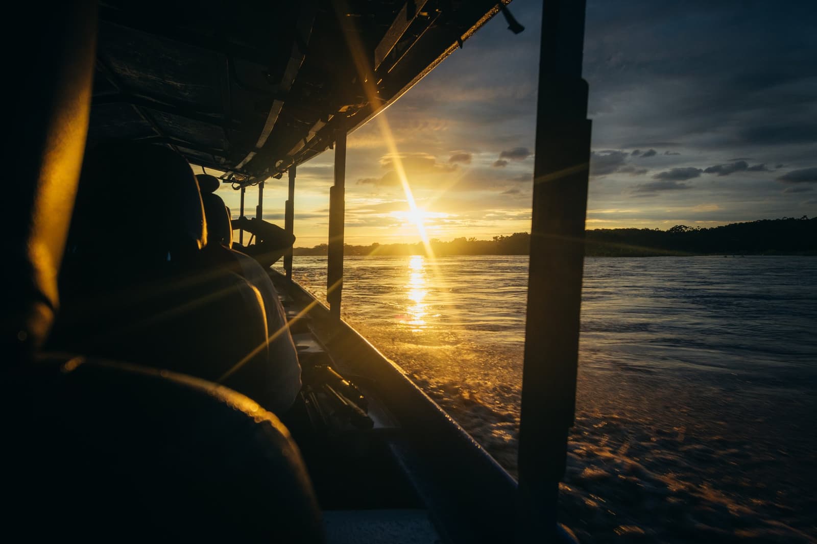 Last look at the river before the forest takes over again. Photo: GABRIEL_S View from the side of a covered river boat on the Tambopata Research Center Peru journey, with the sun setting low over a wide Amazonian river and dark forest silhouetted on the far bank