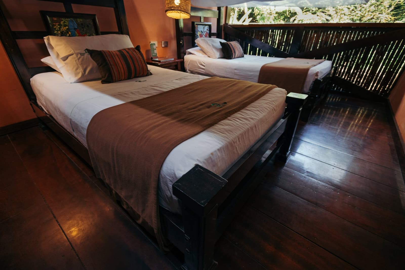 Comfortable beds, forest just past the window. You earned both. Photo: GABRIEL_S Twin-bed room at the Tambopata Research Center Peru lodge, with dark hardwood floors, warm terracotta walls, striped accent pillows, woven pendant lamp, and a bamboo-screened window opening onto the forest canopy