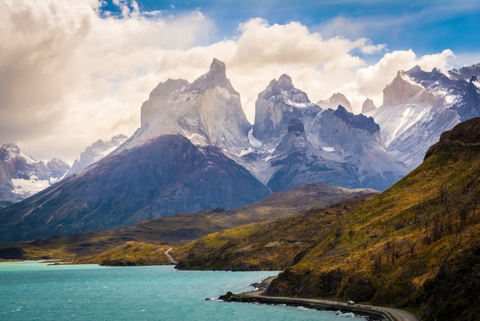 That turquoise lake is not a filter. Patagonia just looks like this. The jagged granite peaks of Los Cuernos rise above a turquoise glacial lake in Torres del Paine, with a winding road hugging the shoreline and green hillsides in the foreground on this torres del paine 4 day all inclusive tour