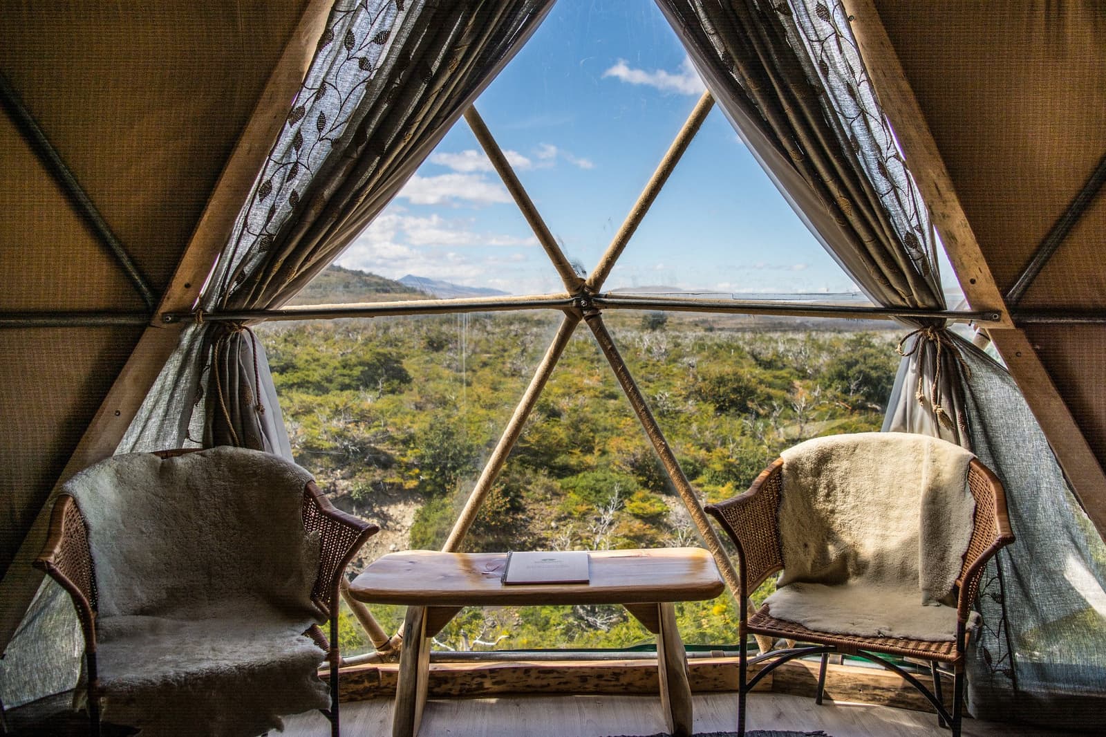 Two chairs, a book on the table, and all of Patagonia waiting just outside. Photo: Suite Dome Interior of a geodesic dome on the torres del paine 4 day all inclusive tour, with two rattan chairs draped in sheepskin throws flanking a rustic wood table, and a large triangular glass panel framing a wide Patagonian scrubland under blue sky