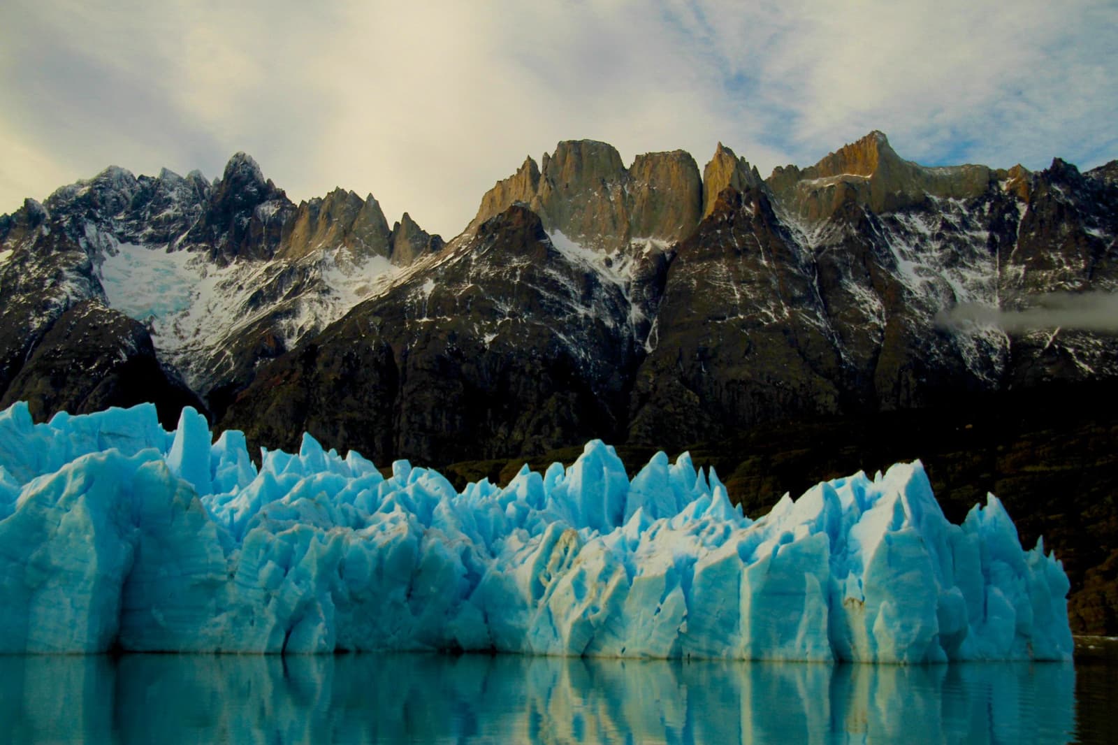 That blue runs deeper than any photo suggests. See it from the boat. Jagged blue ice seracs of Grey Glacier rise from turquoise glacial water on the Torres del Paine W Trek, with snow-dusted granite peaks catching warm light in the background