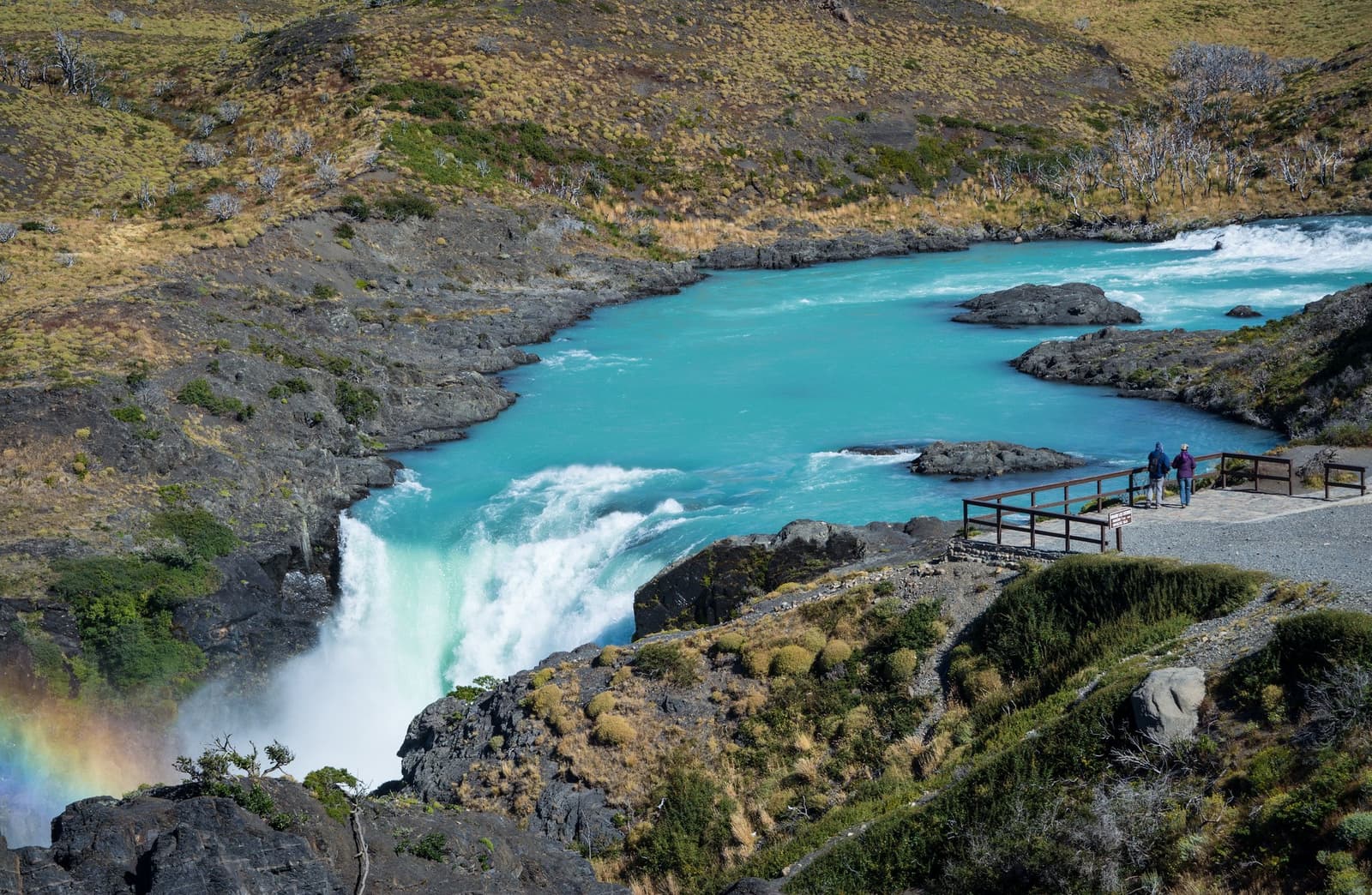 That turquoise is glacier melt, and it really is that colour. Two hikers stand at a wooden viewpoint railing above a powerful waterfall on the Torres del Paine W Trek, looking out over a wide pool of vivid turquoise glacial water framed by rocky slopes and dry Patagonian scrub, with a small rainbow visible in the mist below