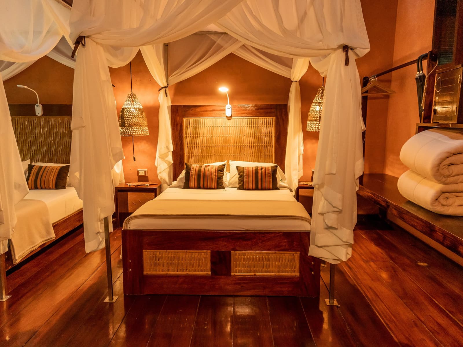 Crisp linen, warm timber, and the jungle humming just outside the open walls. Four-poster canopy bed with white mosquito netting, woven rattan headboard, striped accent pillows, bamboo pendant lamps, and dark hardwood floors inside a Posada Amazonas lodge room