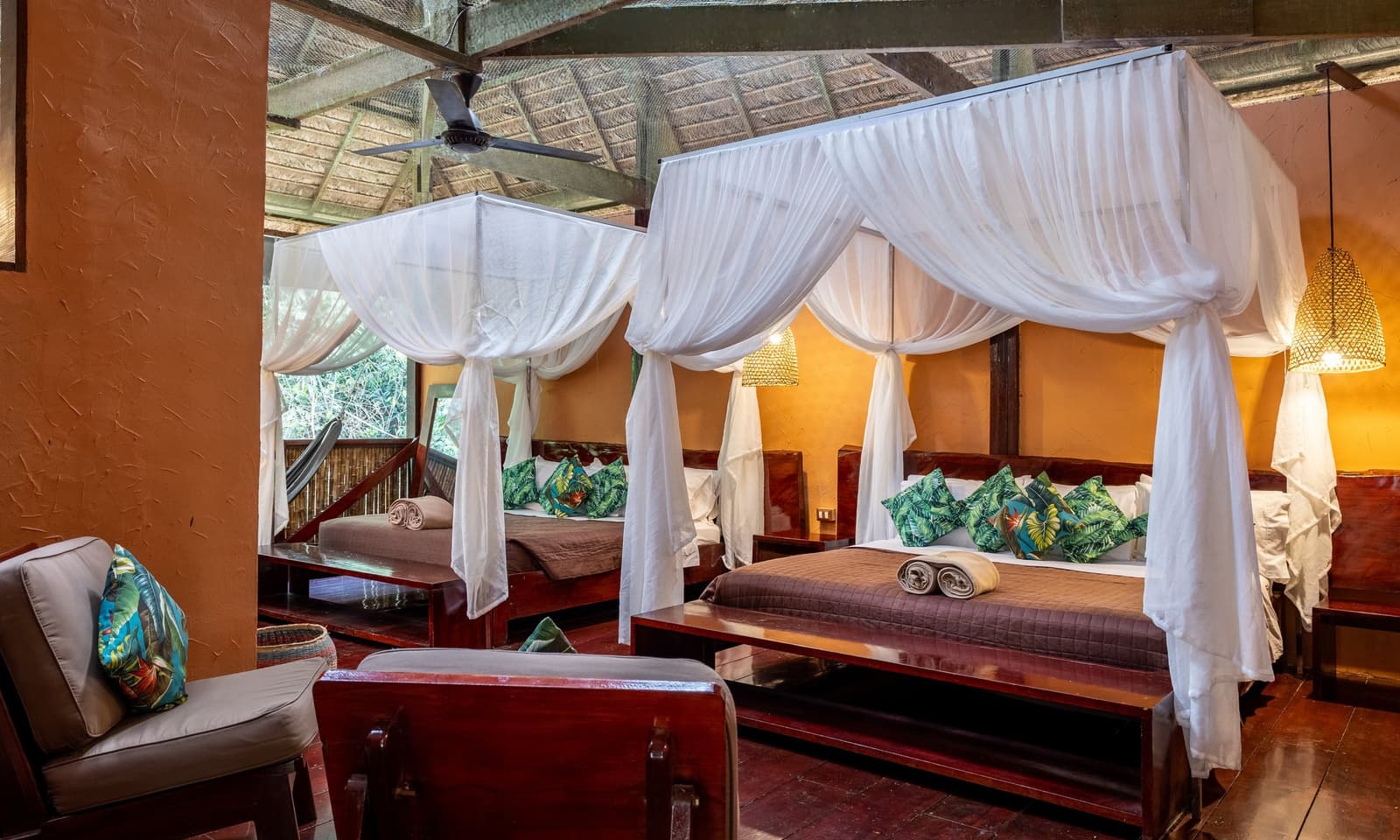 Comfort and the jungle share the same room here, no wall required. Twin four-poster beds with white mosquito net canopies, tropical-print pillows, and warm rattan pendant lighting inside a thatched-roof room at Posada Amazonas lodge, with jungle views through the open wall