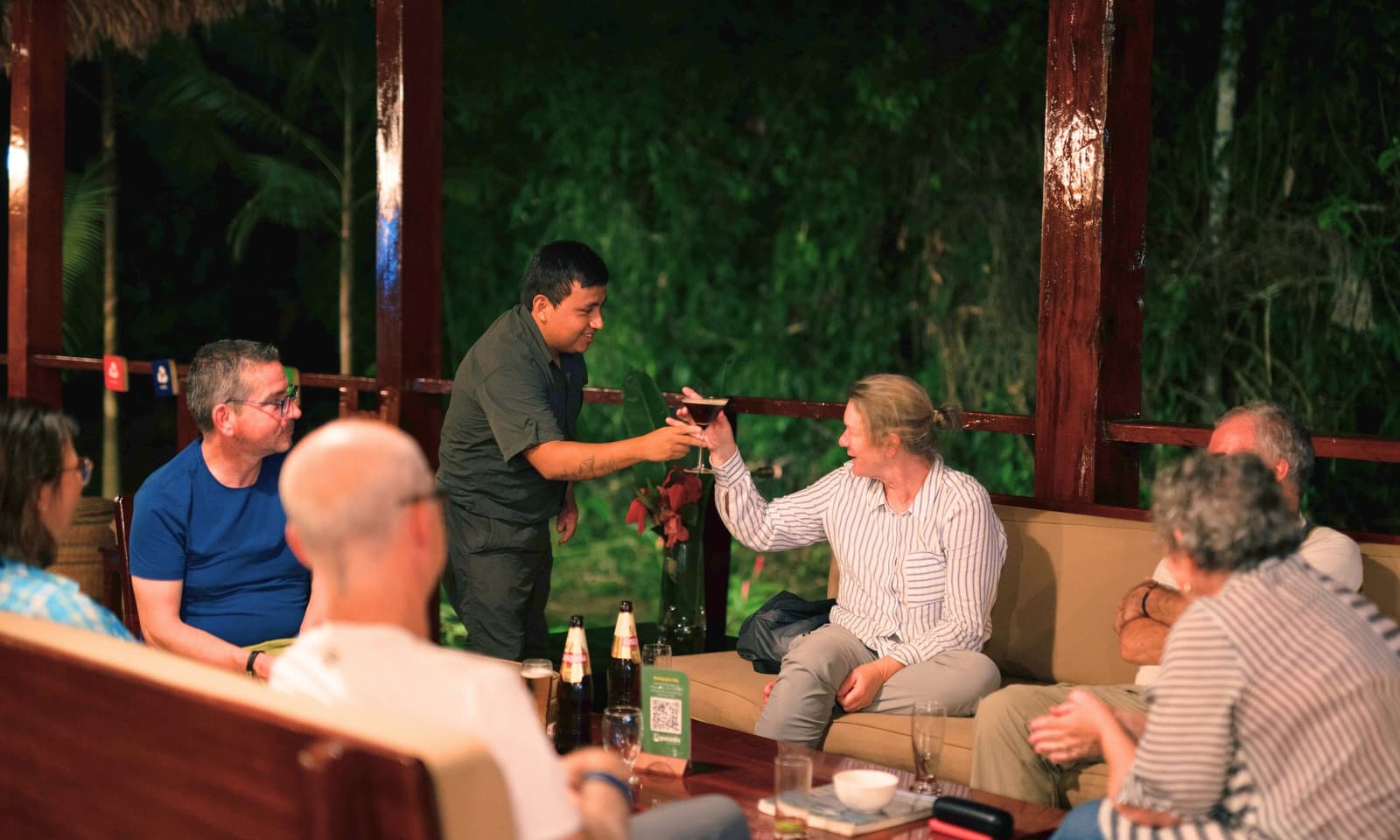 Good drinks, good company, and the jungle just a few feet away. A staff member at a jungle lodge hands a cocktail to a smiling guest during an amazon tour from puerto maldonado, as a small group relaxes in an open-air lounge surrounded by nighttime rainforest