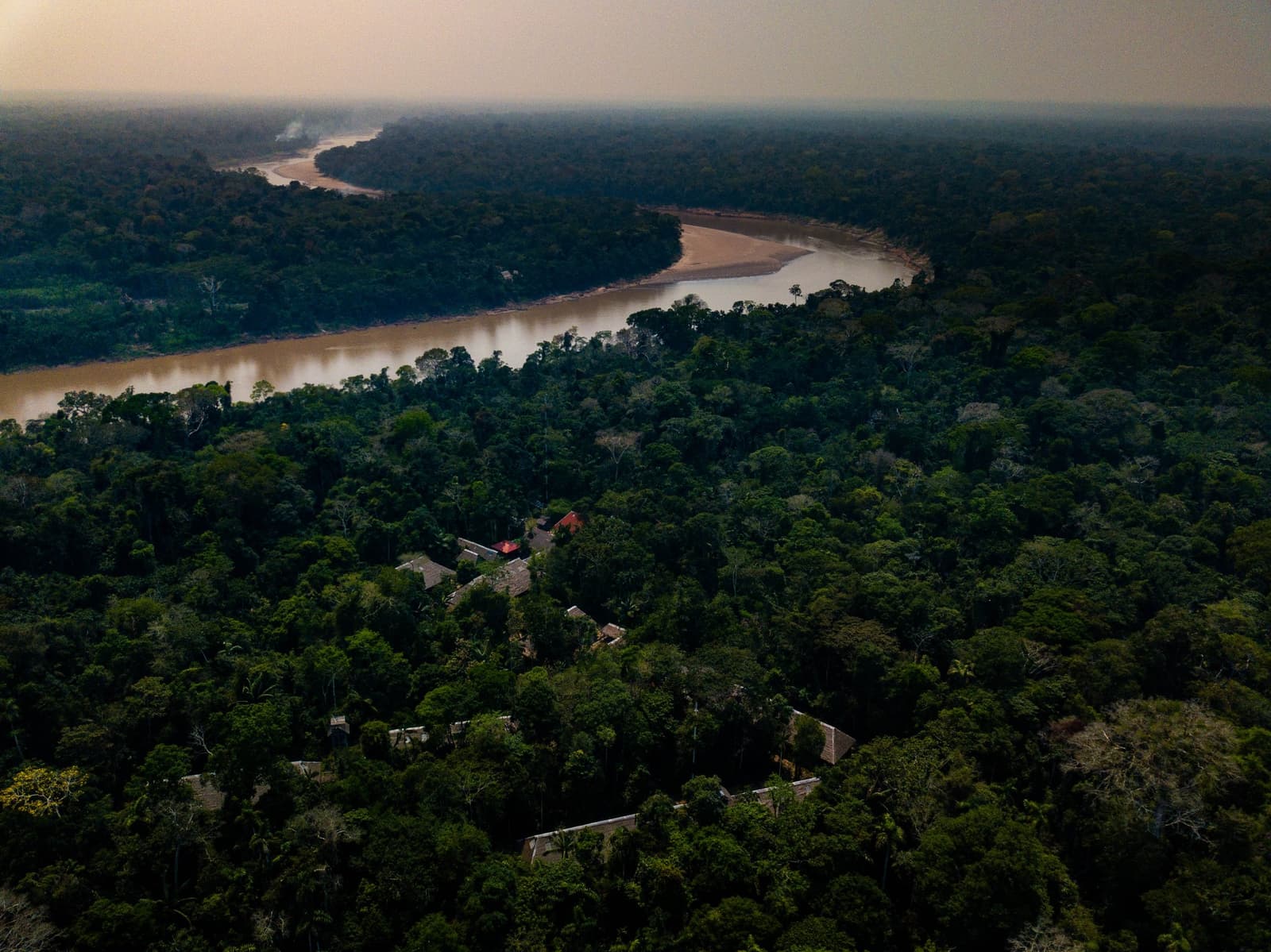 Tucked into the canopy, the lodge is easier to find by boat than by eye. Aerial view of a rainforest lodge nestled among dense Amazon canopy alongside a wide, sandy-banked river winding into the hazy horizon, seen on an amazon tour from puerto maldonado
