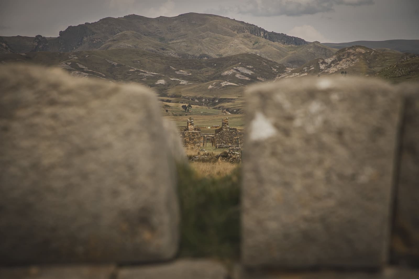 Pedras emoldurando pedras. Os Andes sempre souberam sobrepor a história. Vista através de uma abertura em blocos de pedra desgastados na trilha Ancascocha, enquadrando ruínas distantes e colinas andinas onduladas sob um céu nublado