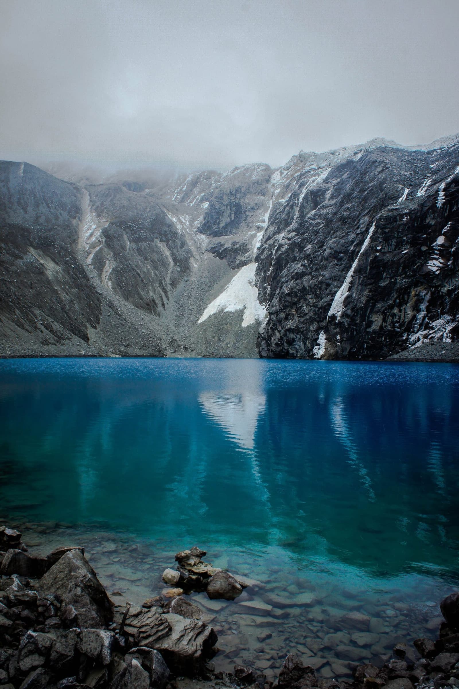 Água gelada, cor impossível, e um glaciar que parece quase ao alcance da mão. Um lago glacial de turquesa intenso e azul profundo reflete penhascos rochosos íngremes com listras de neve sob um céu encoberto no trek Ancascocha, com pedras lisas visíveis através da água clara e rasa do primeiro plano.