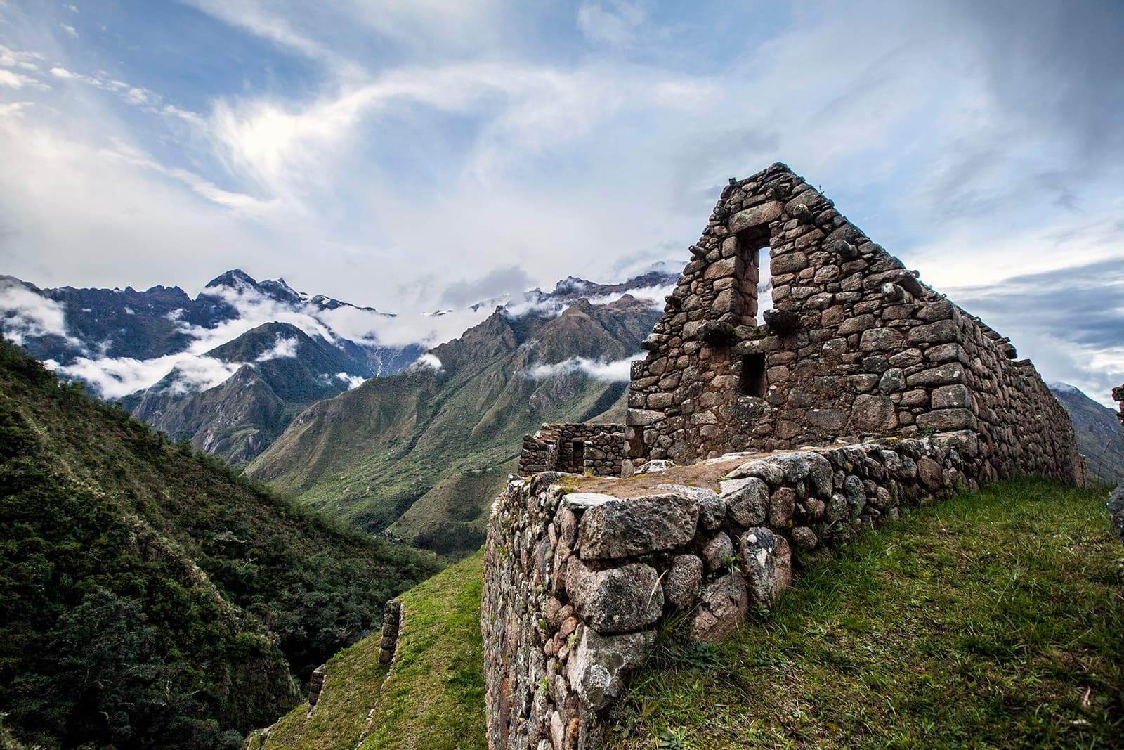 Stone walls that have outlasted every empire, still holding their ground up here. Roofless Inca stone structure perched on a high ridge during an andes trek, with cloud-draped Andean peaks and deep green valleys stretching into the distance