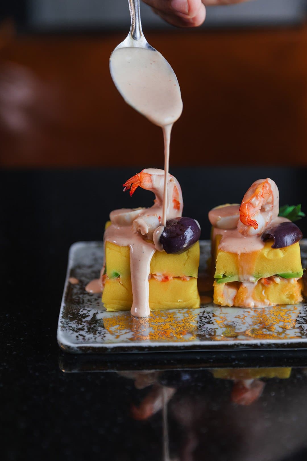 Lunch on a river that moves at its own pace, the kitchen keeps up just fine. Photo: Yvonne Kreuzmayr A hand drizzles creamy pink sauce from a spoon over two plated causa towers topped with shrimp and black olives on the Aria Amazon cruise, served on a dark ceramic dish against a black surface