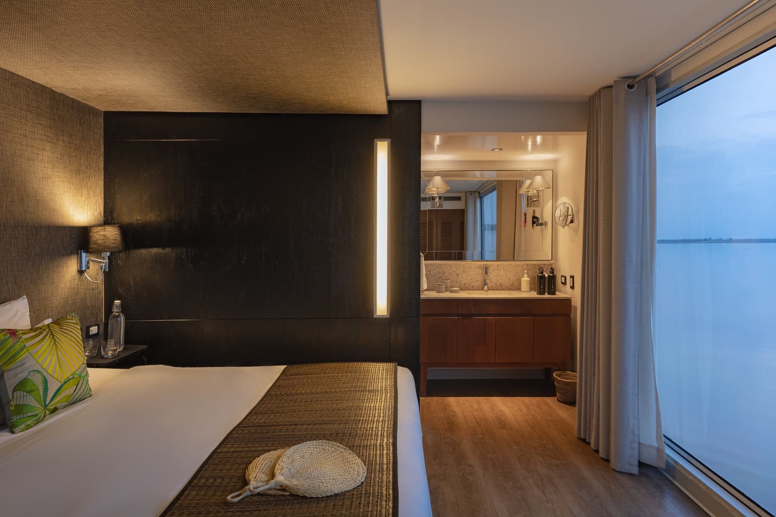 Warm light inside, a wide river outside, and nothing urgent on the schedule. Photo: Francois Moreau A well-appointed cabin on the Aria Amazon cruise features a double bed with tropical print pillows, a woven fan resting on the bed runner, warm wall sconces, an open en-suite bathroom with a wood-panelled vanity, and floor-to-ceiling windows looking out over a wide river at dusk.
