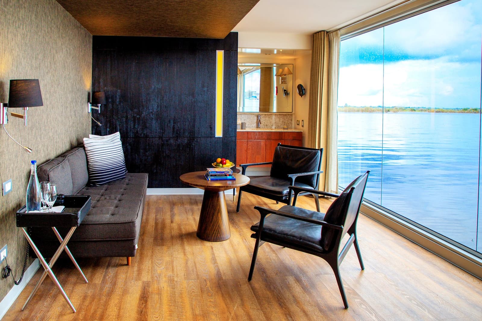 Floor-to-ceiling glass, leather chairs, and the river just there, your living room has a decent view. Photo: Aqua Expeditions Elegantly furnished suite on the Aria Amazon cruise featuring two leather armchairs, a round walnut coffee table with a fruit bowl, a tufted gray sofa, and floor-to-ceiling glass windows overlooking the calm open river