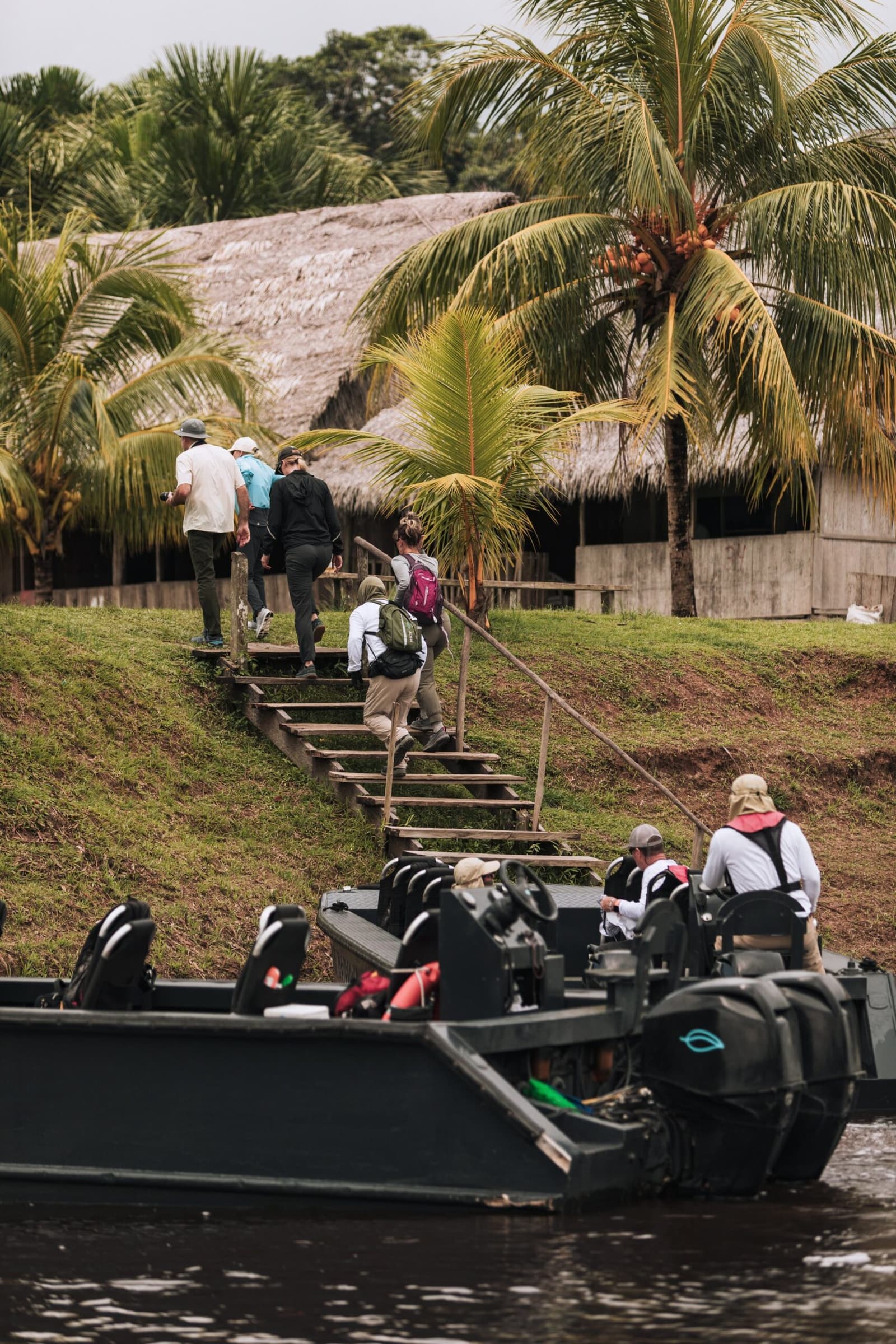 Step off the skiff, climb the bank, and the village is yours to explore. Photo: Yvonne Kreuzmayr Guests on an aria amazon cruise climb wooden riverside steps toward a thatched-roof village building, while two motorized skiffs wait at the water's edge beneath coconut palms