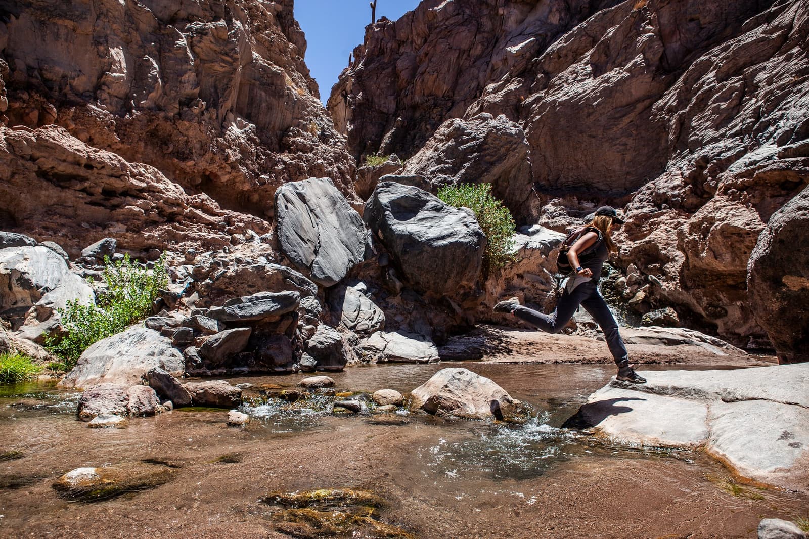 Botas secas, buen propósito. La quebrada tiene sus propios planes. Una senderista con mochila salta sobre un arroyo poco profundo en rocas planas dentro de un estrecho cañón desértico con imponentes paredes de arenito durante una expedición de camping en el desierto de Atacama
