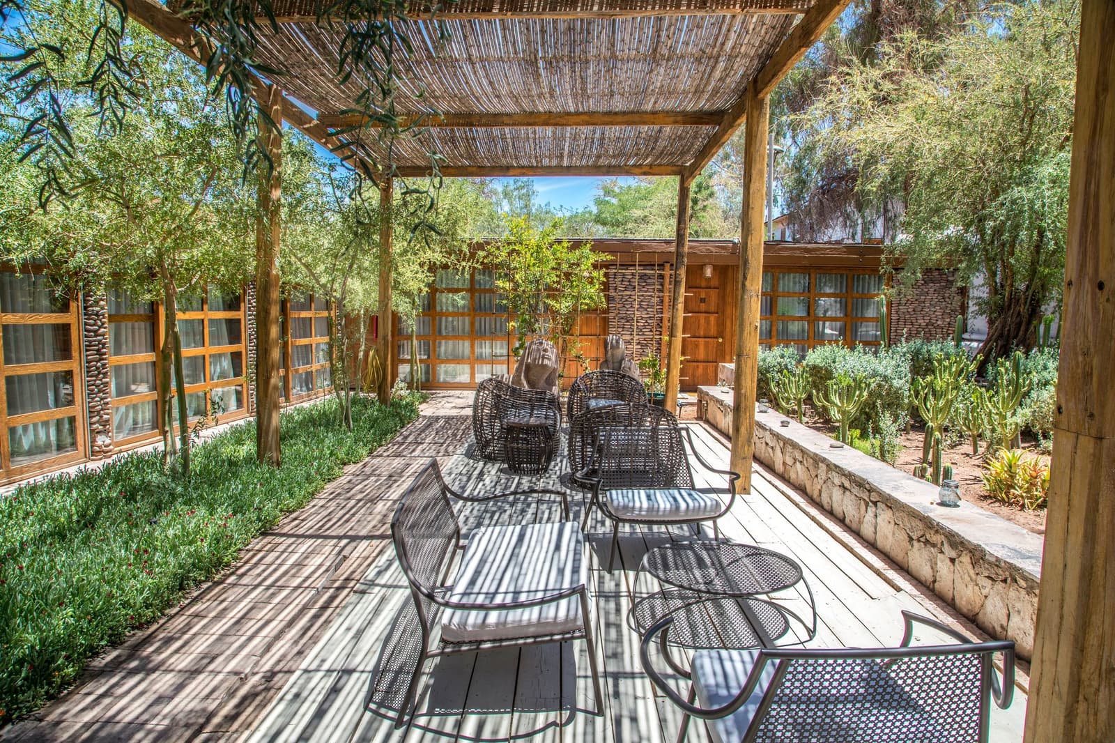 Almuerzo en el jardín, hamacas a mano, el desierto puede esperar. Photo: LUIS HERNAN HERREROS INFANTE Terraza de madera sombreada con sillones de ratán y sillas metálicas bajo una pérgola de caña, rodeada de cactus, arbustos y construcciones de piedra de un lodge en San Pedro de Atacama