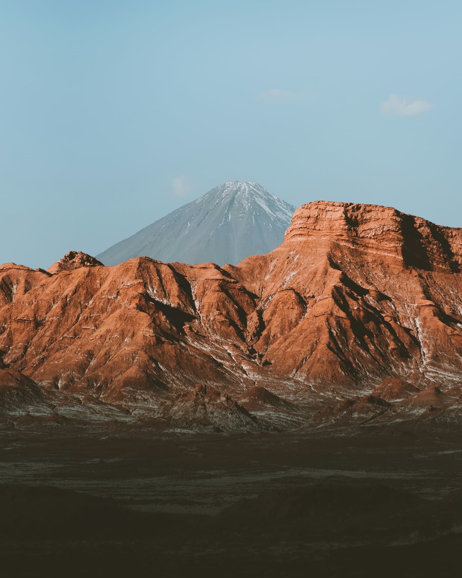 Roca roja en primer plano, volcán al fondo, el Atacama habla por sí solo. Formaciones rocosas rojas en capas en el desierto de Atacama con un volcán cónico nevado elevándose detrás bajo un cielo azul pálido
