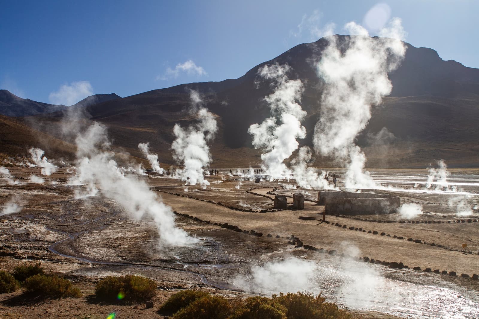 The ground here is essentially boiling. Bring a jacket. Photo: Tatio Campo Geotermico Dozens of geothermal steam vents rising from an arid high-altitude plain at El Tatio on this atacama desert tour package, with a dark volcanic mountain and clear blue sky in the background and visitors walking among the fumaroles