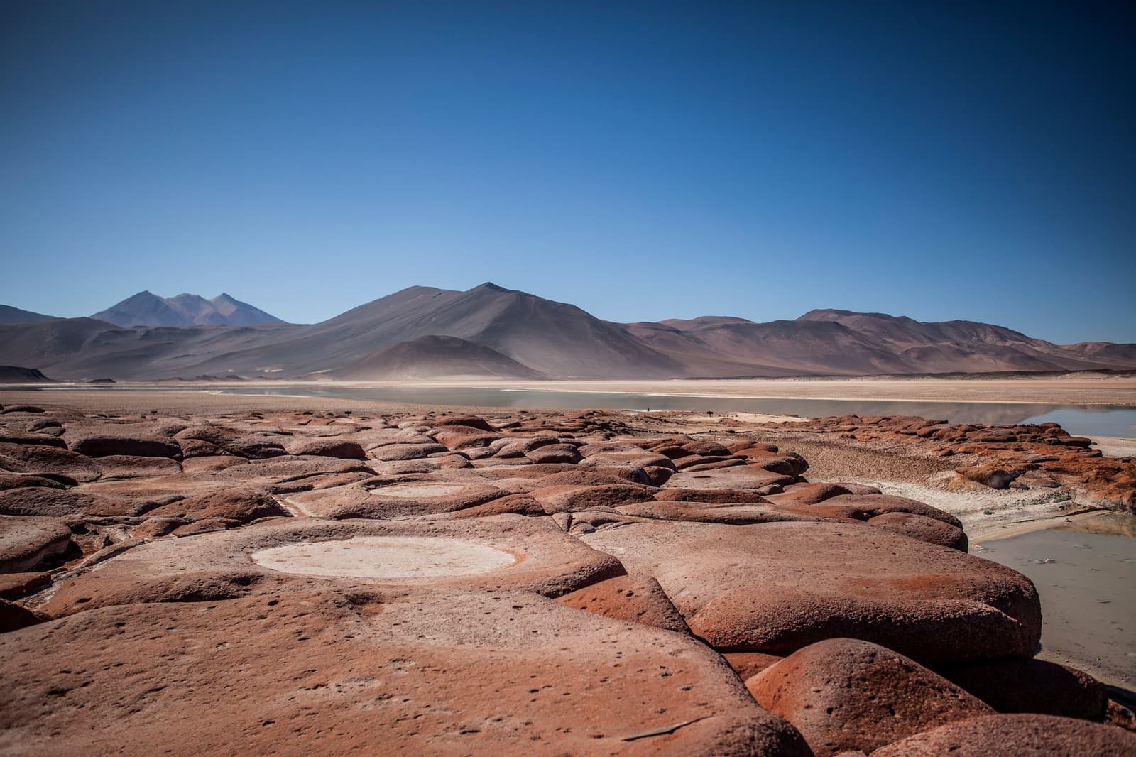 Red rocks, a ghost-blue lagoon, and volcanoes that forgot to hurry. Photo: Piedras Rojas Rounded red rock formations at Piedras Rojas in the Atacama desert tour package, with a pale turquoise salt flat lagoon and bare volcanic mountains under a deep blue sky