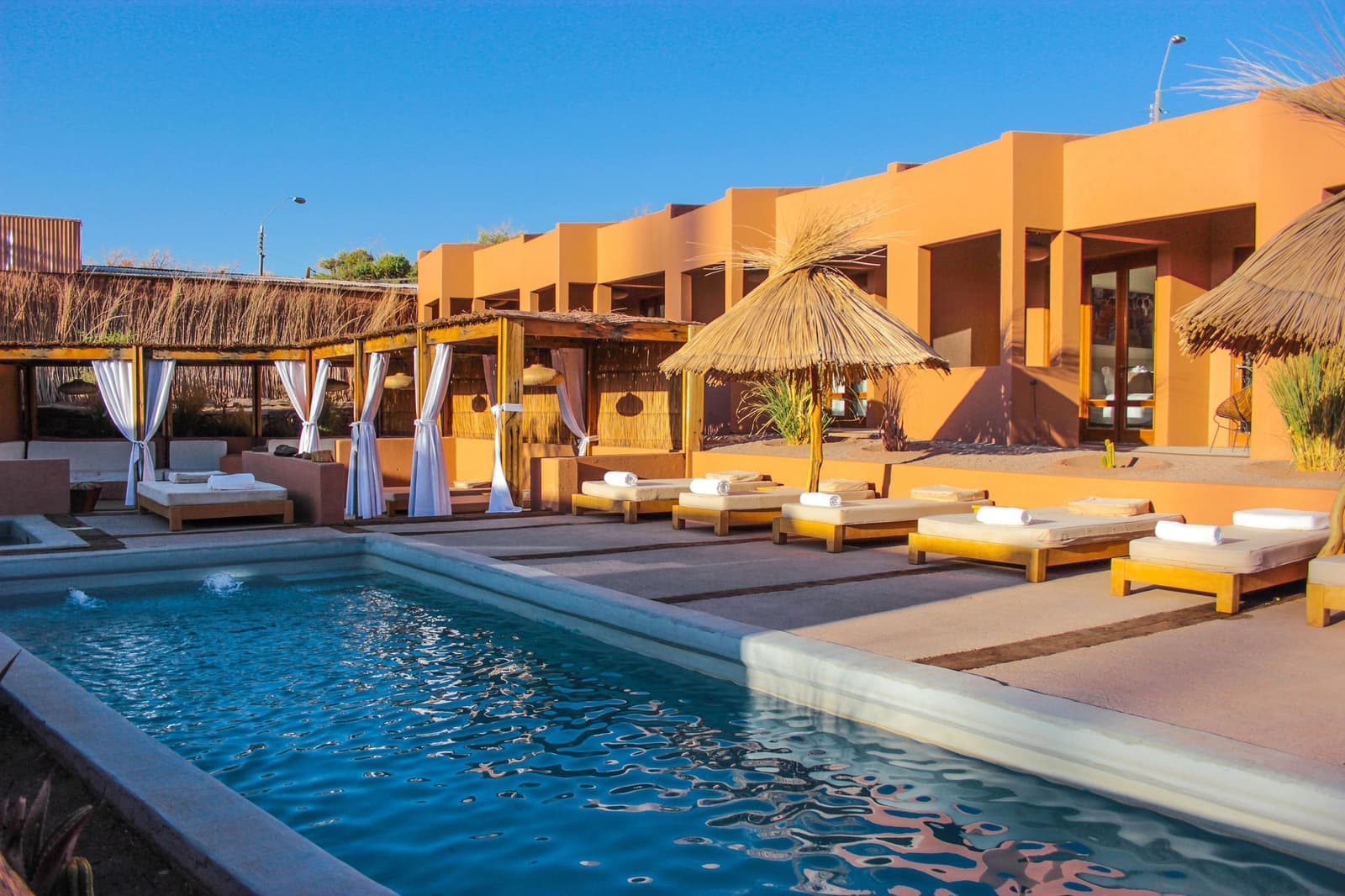 Cold water, warm walls, and the whole afternoon still ahead of you. The outdoor pool area of a desert lodge included in an Atacama Desert tour package, with wooden sun loungers topped with rolled white towels, thatched parasols, white-curtained cabanas, and warm terracotta-toned walls under a deep blue sky