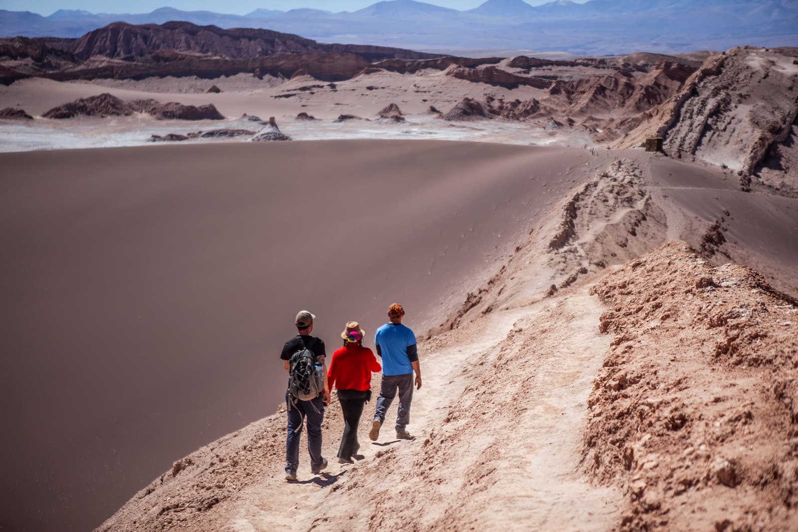 Three people, one ridge, and a landscape that makes you recalibrate your sense of scale. Three hikers walk along a narrow ridge above the Great Dune in Valle de la Luna during an Atacama Desert tour package, with sculpted salt formations and distant Andean peaks stretching to the horizon