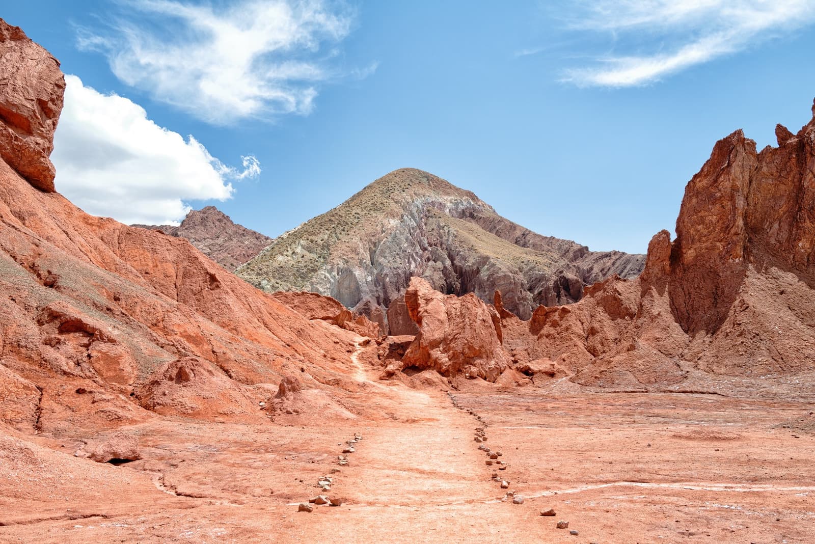 Stenen als gids, de berg als eindbestemming. Een met stenen gemarkeerd pad leidt door geërodeerde rode rotsformaties tijdens de Atacama-woestijntrek, met een kegelvormige groen-oker berg onder een blauwe hemel