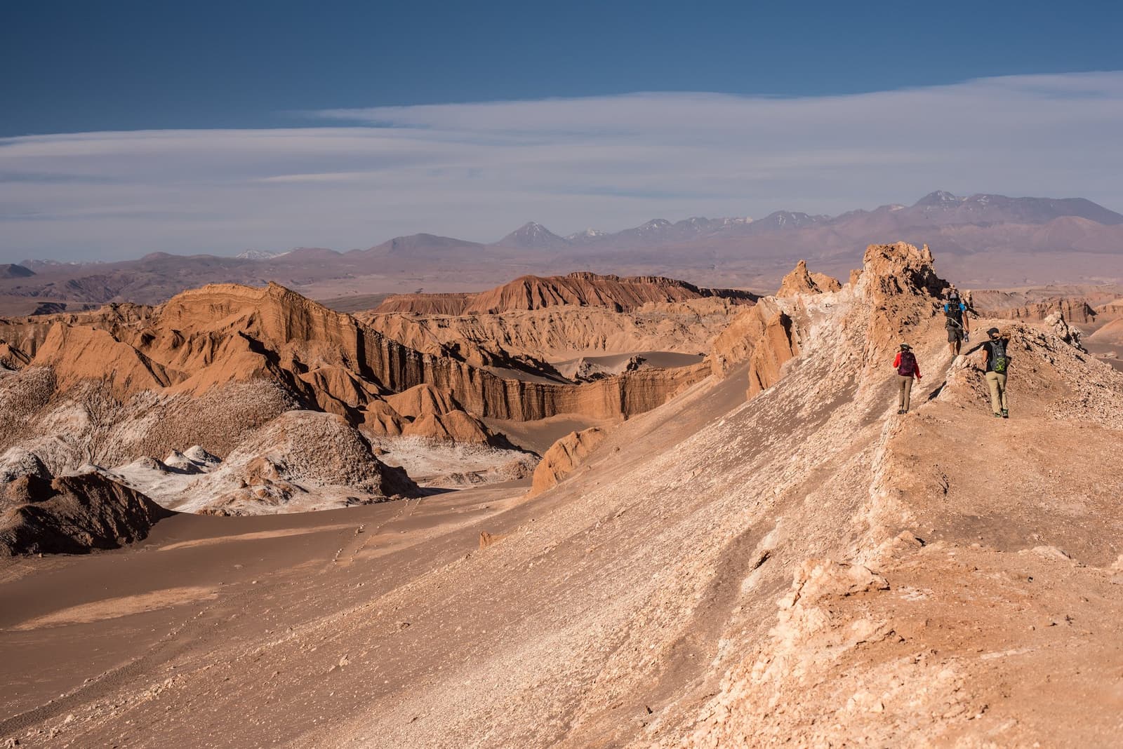 Drie rugzakken, één smalle richel, en heel veel uitzicht. Drie wandelaars met rugzakken lopen langs een smalle zout-en-rotsrug tijdens een Atacama desert trek, met geërodeerde okergele badlands, witte zoutvlakten en besneeuwde Andeskraters die zich uitstrekken tot de horizon onder een heldere blauwe lucht.