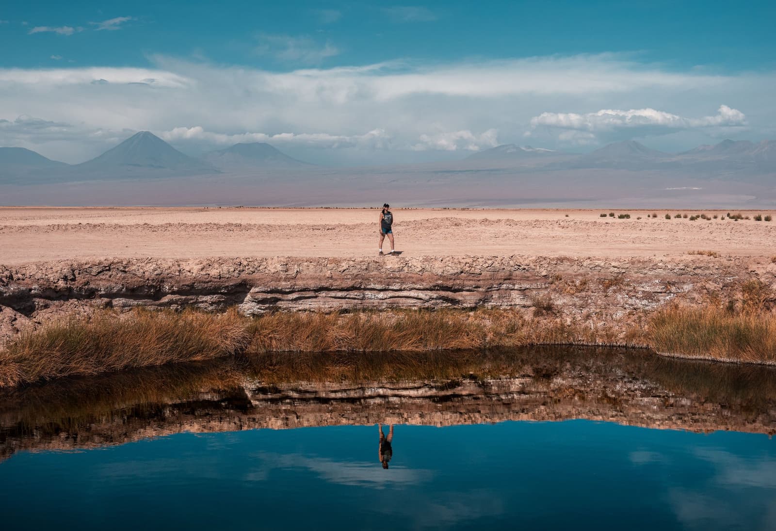 Jij en je spiegelbeeld, allebei even verrast door de stilte hier. Een eenzame wandelaar staat op een zoutkorst richel boven een spiegelstille lagune tijdens een Atacama woestijn trek, volledig weerspiegeld in het blauwe water met vulkanen boven de vlakte op de achtergrond