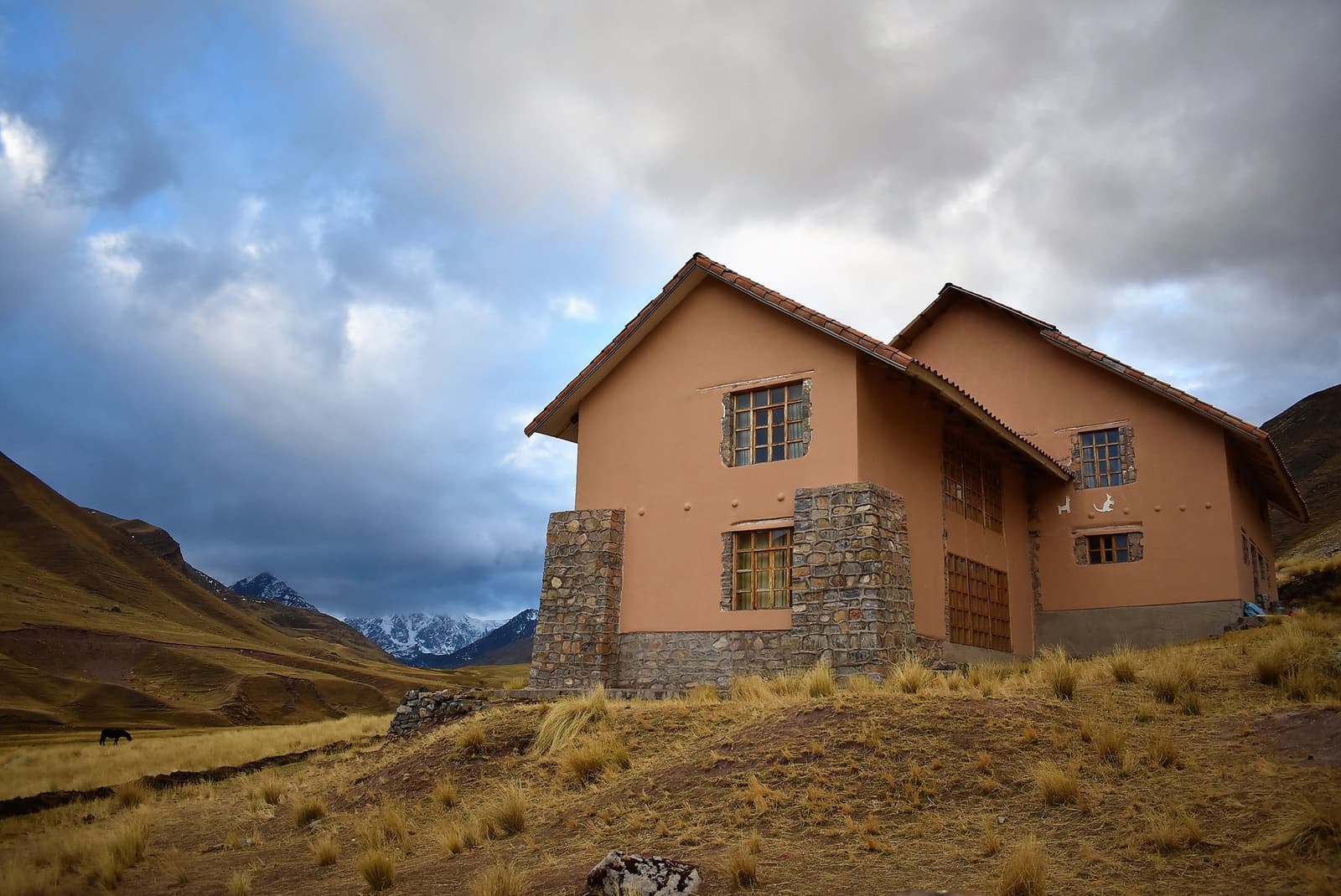 Stone and plaster, built to last at altitude. Your bed for tonight earned. Photo: Two-storey terracotta-rendered lodge with stone base and tiled roof set on high-altitude grassland, part of the Ausangate Lodge Trek route, with snow-capped Andean peaks visible through clouds in the background and a horse grazing in the valley below