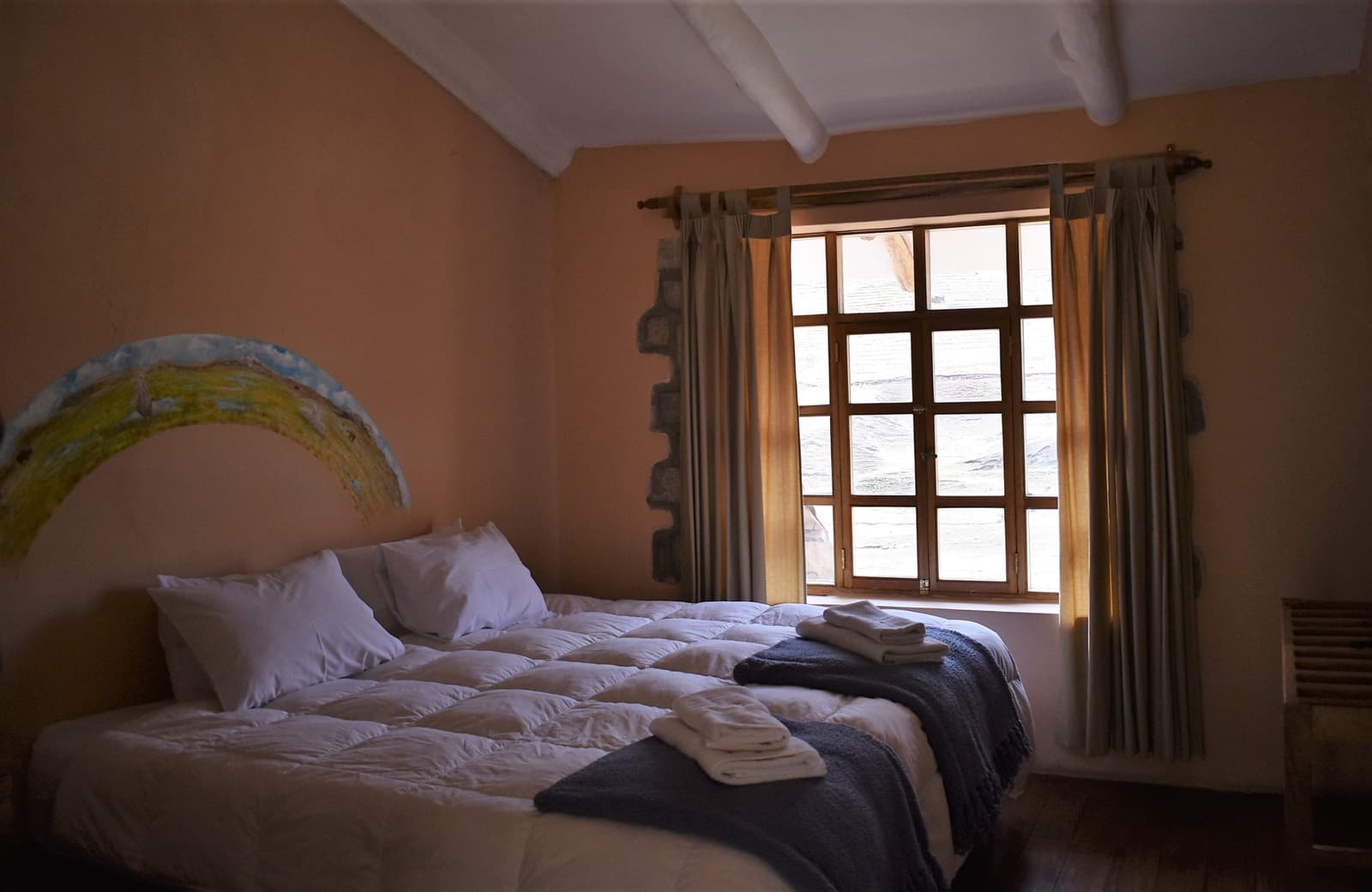 Folded towels, a thick duvet, and a window that frames the Andes. Not bad for the high mountains. Photo: A double bed with white thermal duvet and folded towels inside a stone-walled lodge room on the Ausangate Lodge Trek, with a wooden-framed window and warm terracotta walls